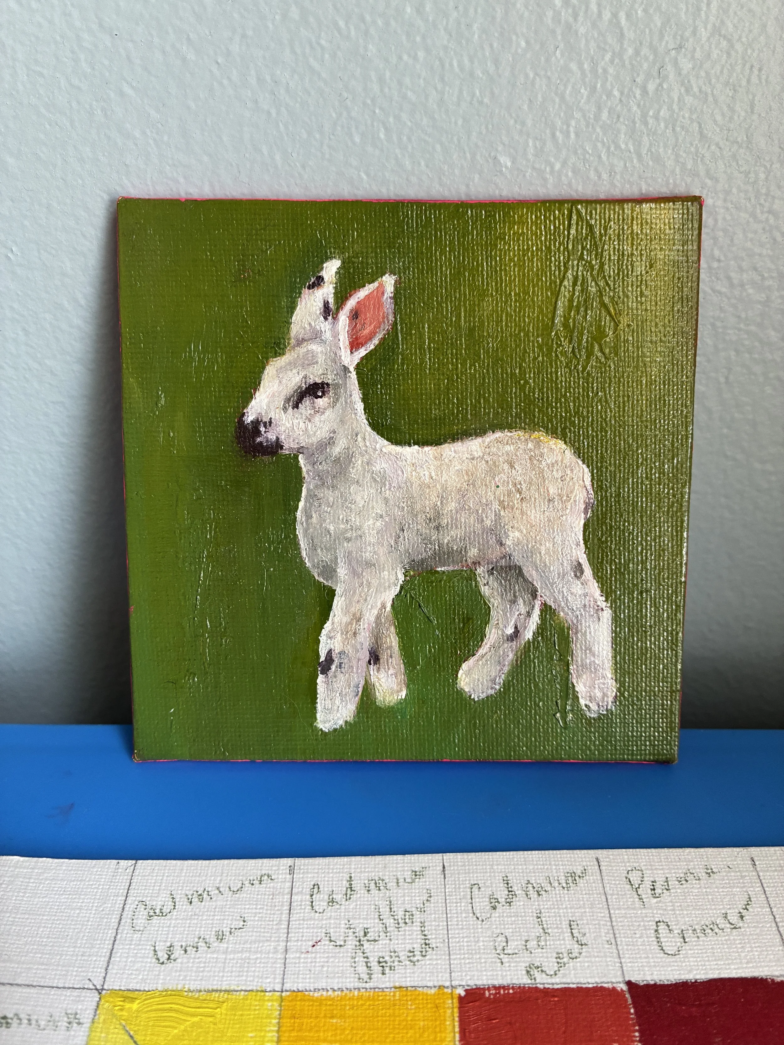 A painting of a white lamb with black spots on a green and yellow background, leaning against a blue wall.