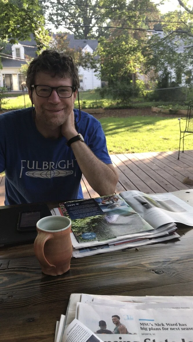 White man with brown hair and glasses in blue shirt with "Fulbright" written on it in white sitting in front of a green lawn