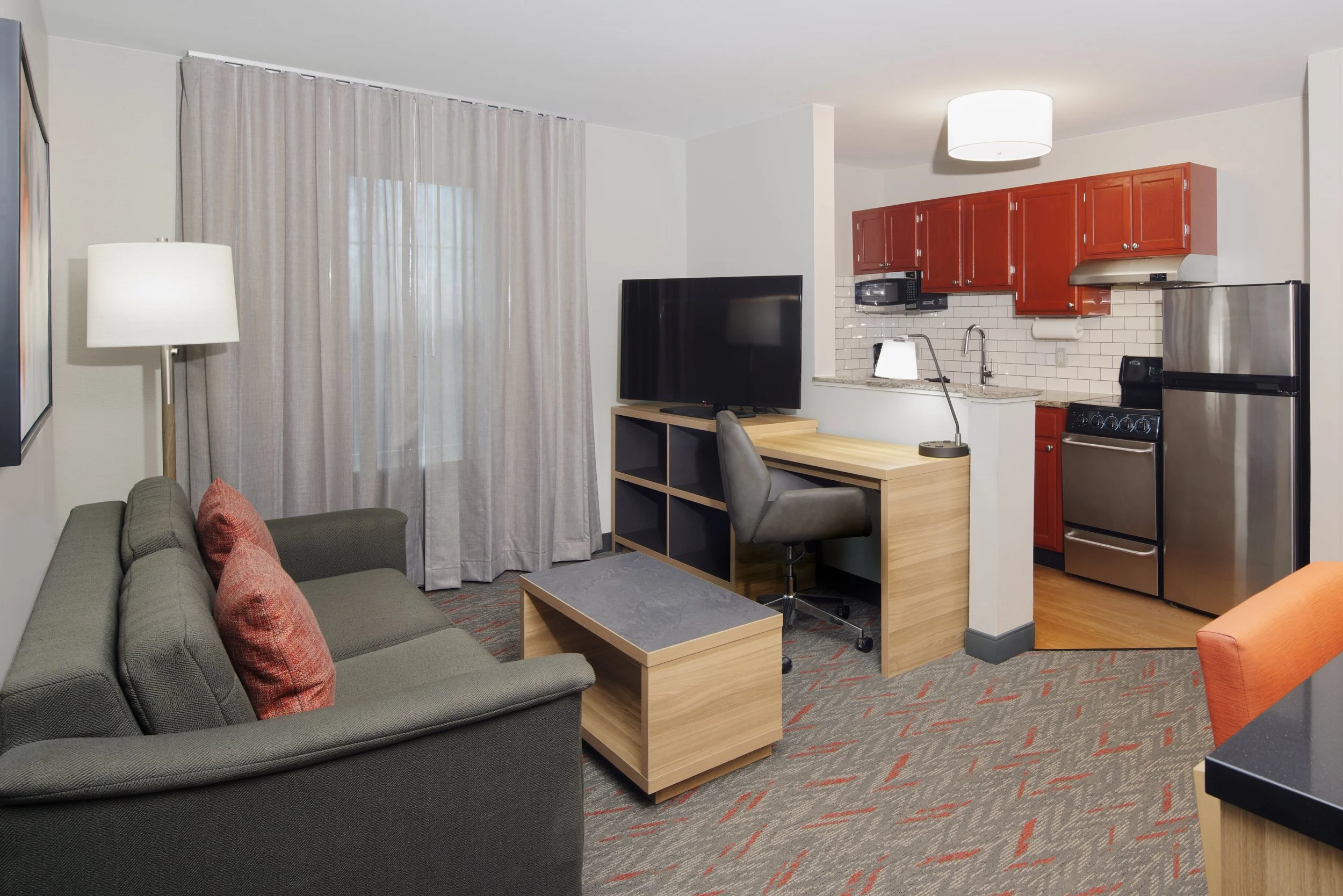 Living area with a green sofa, red pillows, floor lamp, wall art, flat-screen TV, and a work desk with a chair. Kitchen area with red cabinets, microwave, stovetop, refrigerator, and white tile backsplash.