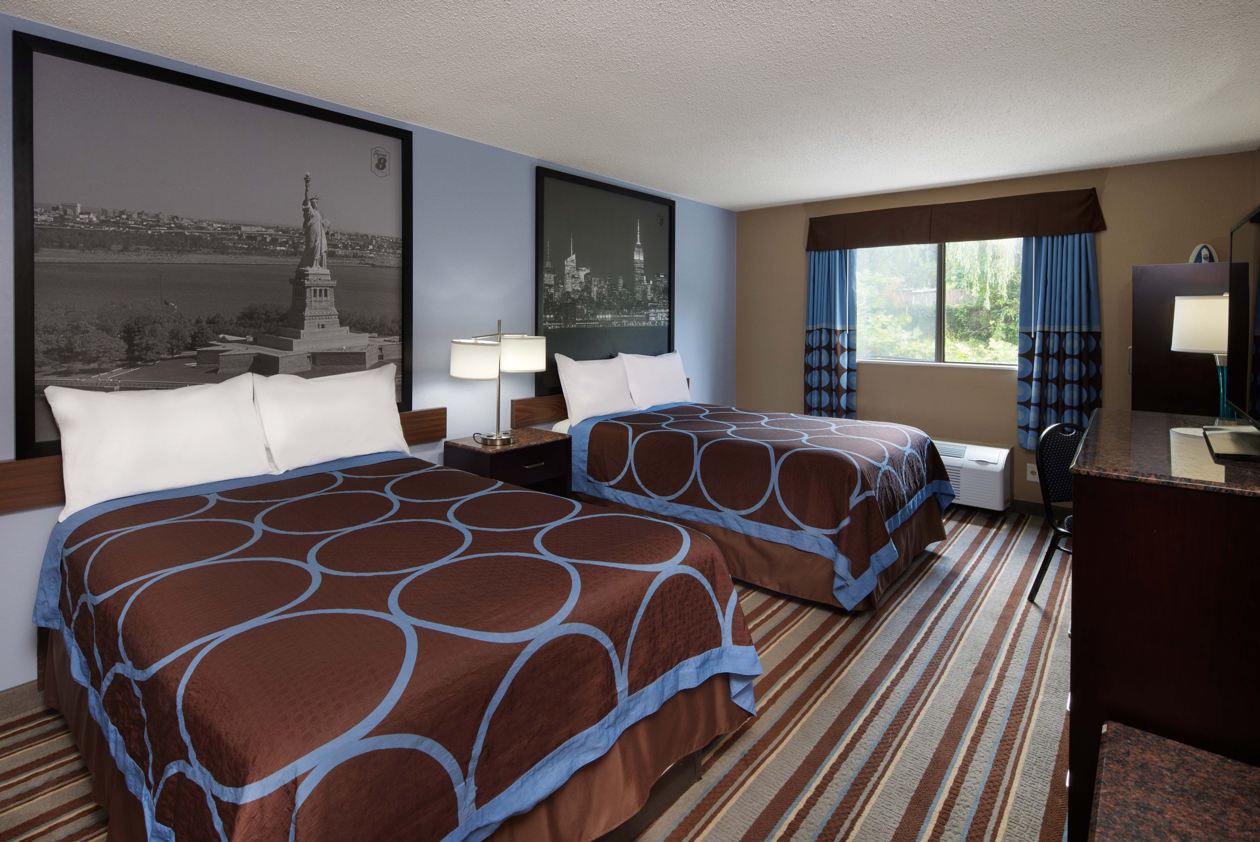 Hotel room with two queen beds with brown and blue patterned bedding, large black and white cityscape pictures above each bed, a window with blue and brown curtains, a desk with a television, and striped carpet.
