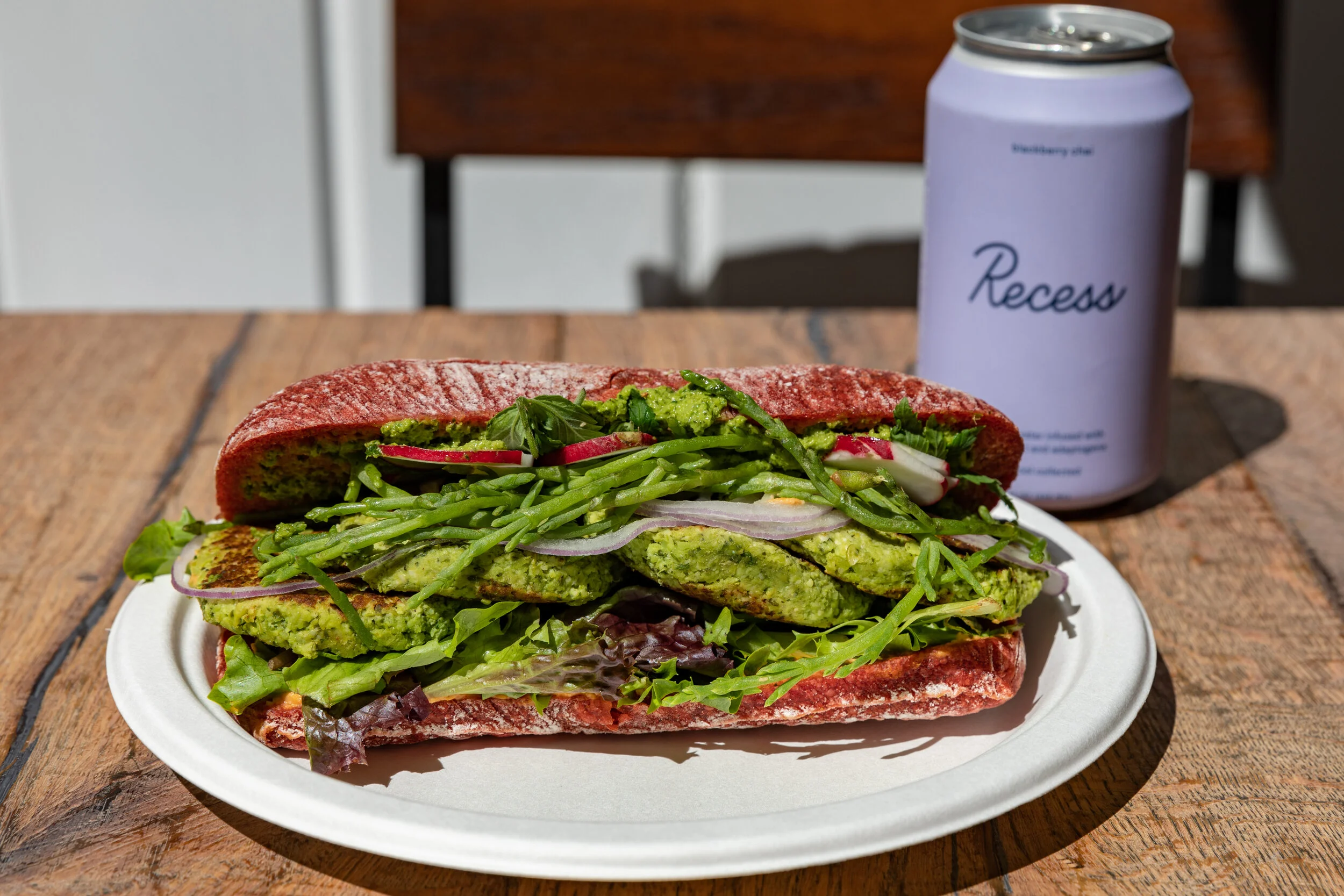 Vegan sandwich with green falafel, cucumber, radish, leafy greens, and herbs on a red bread roll, served on a white plate with a can of Recess sparkling water in the background on a wooden table.