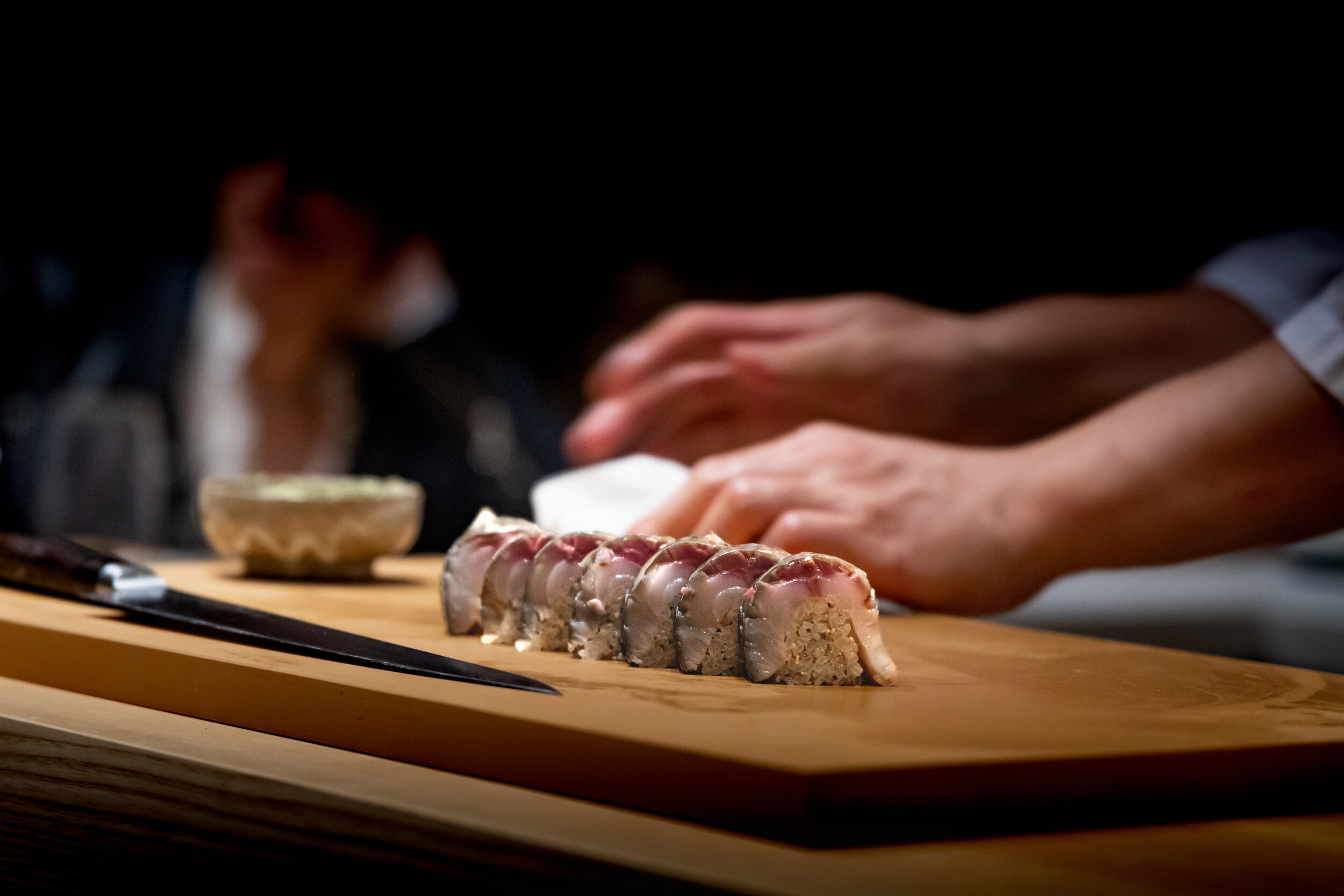 Chef preparing sushi rolls with sliced fish on a wooden cutting board, with a blurred person in the background.