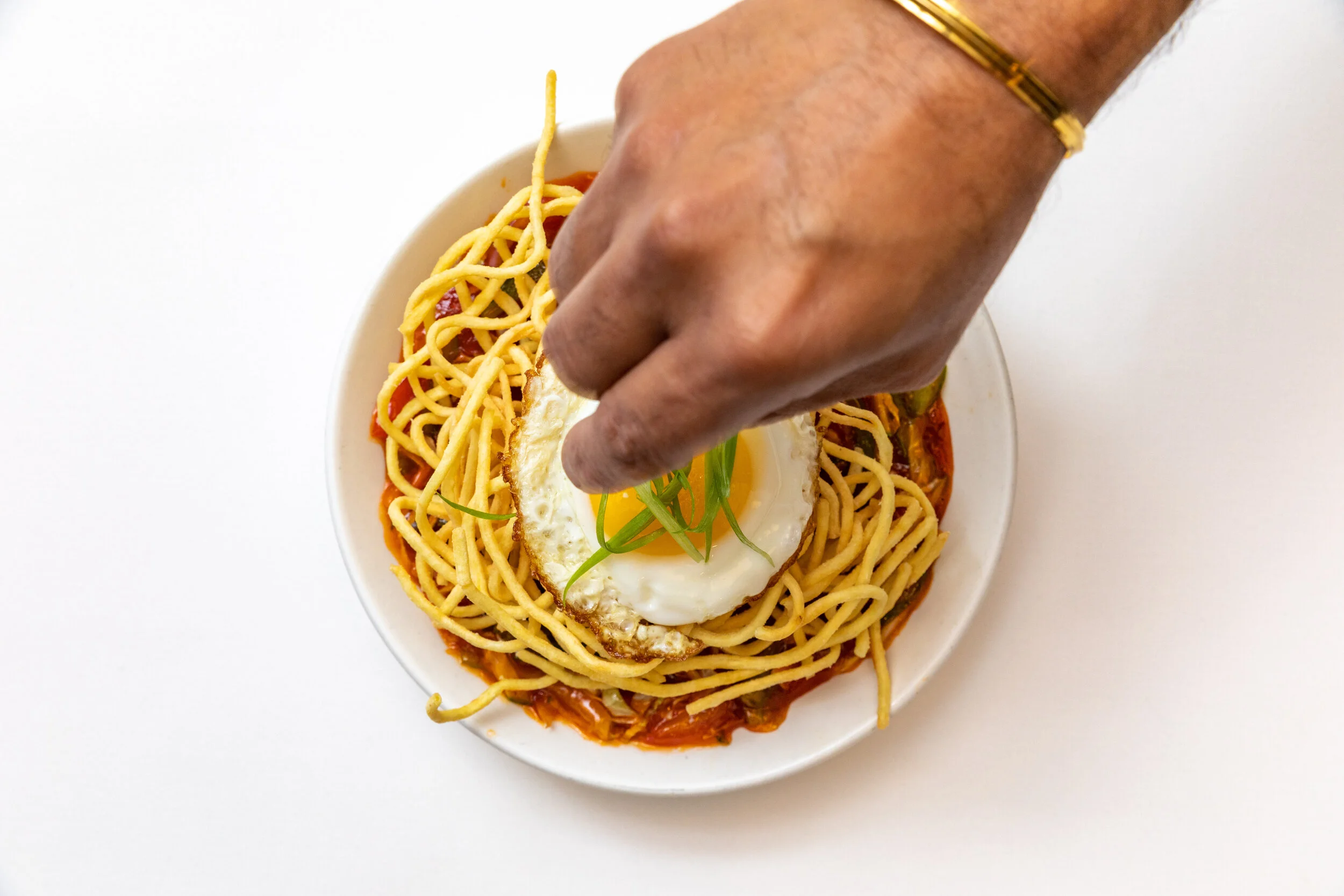 Hand placing a fried egg on top of spaghetti noodles and tomato sauce in a white bowl, garnished with green herbs.