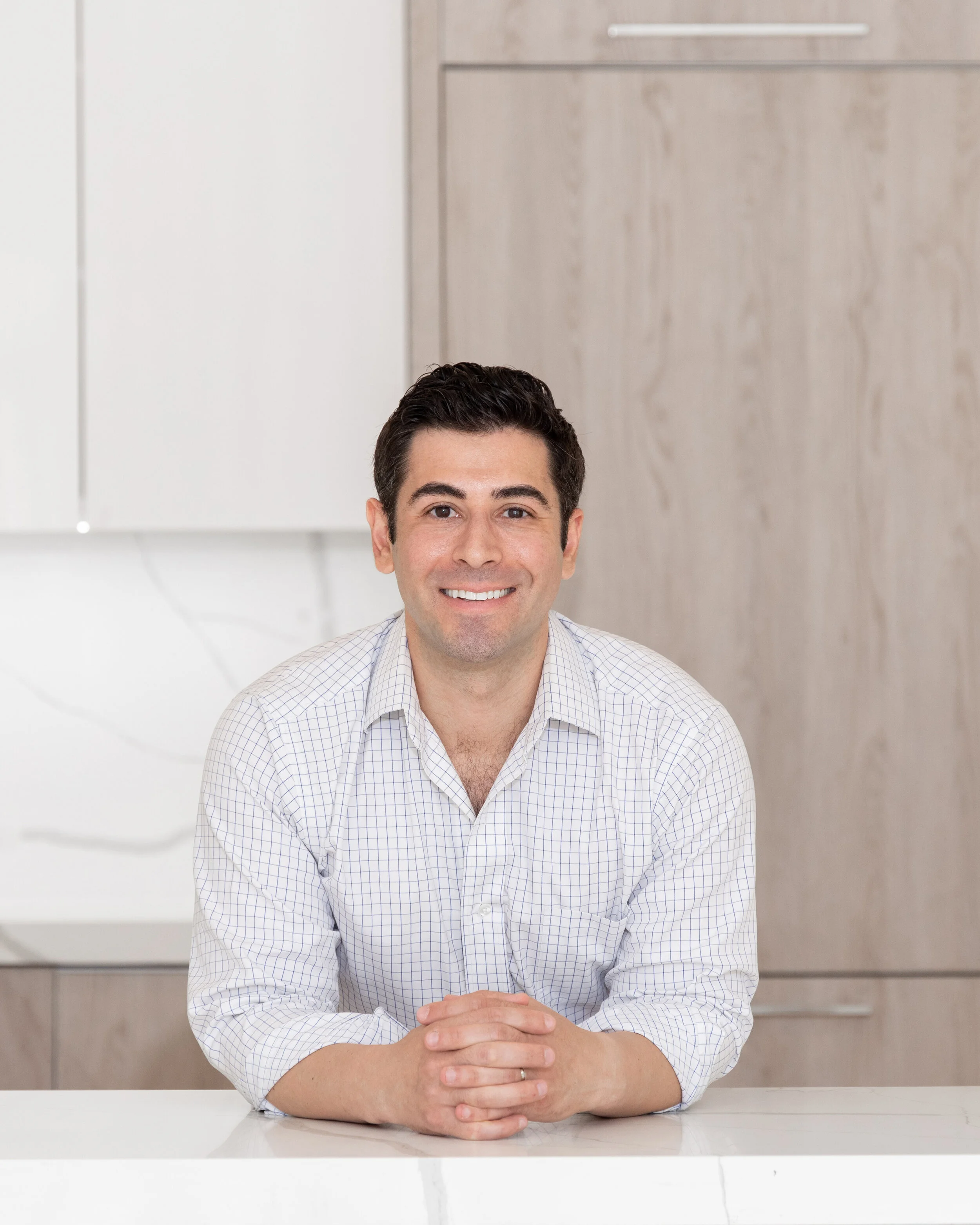 A man with dark hair and a checked button-up shirt sitting at a white table, smiling, with a background of light-colored cabinets.