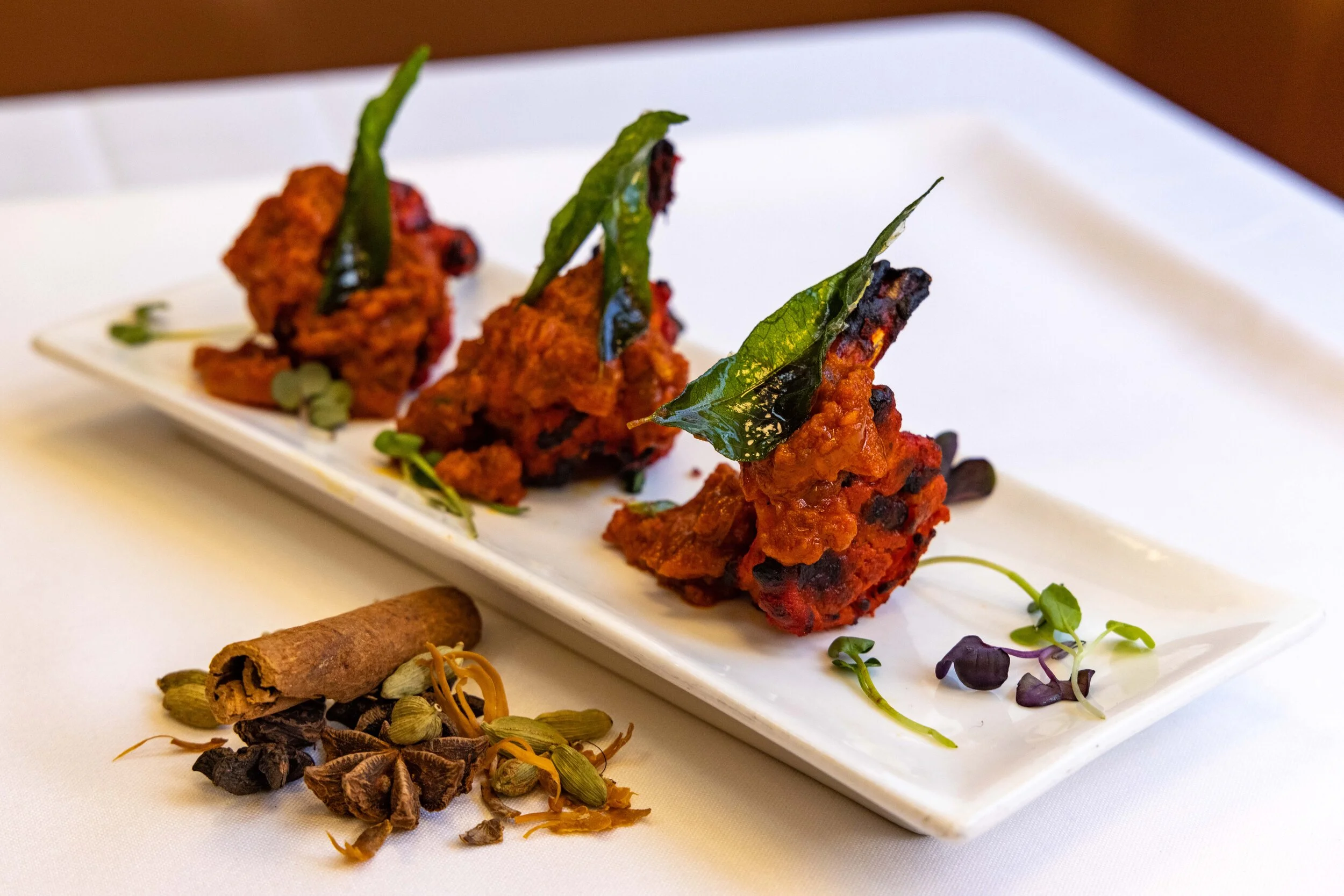 Three fried chicken drumsticks with chili, topped with green leaves, served on a rectangular white plate, garnished with microgreens, with a cinnamon stick and assorted spices on the side.