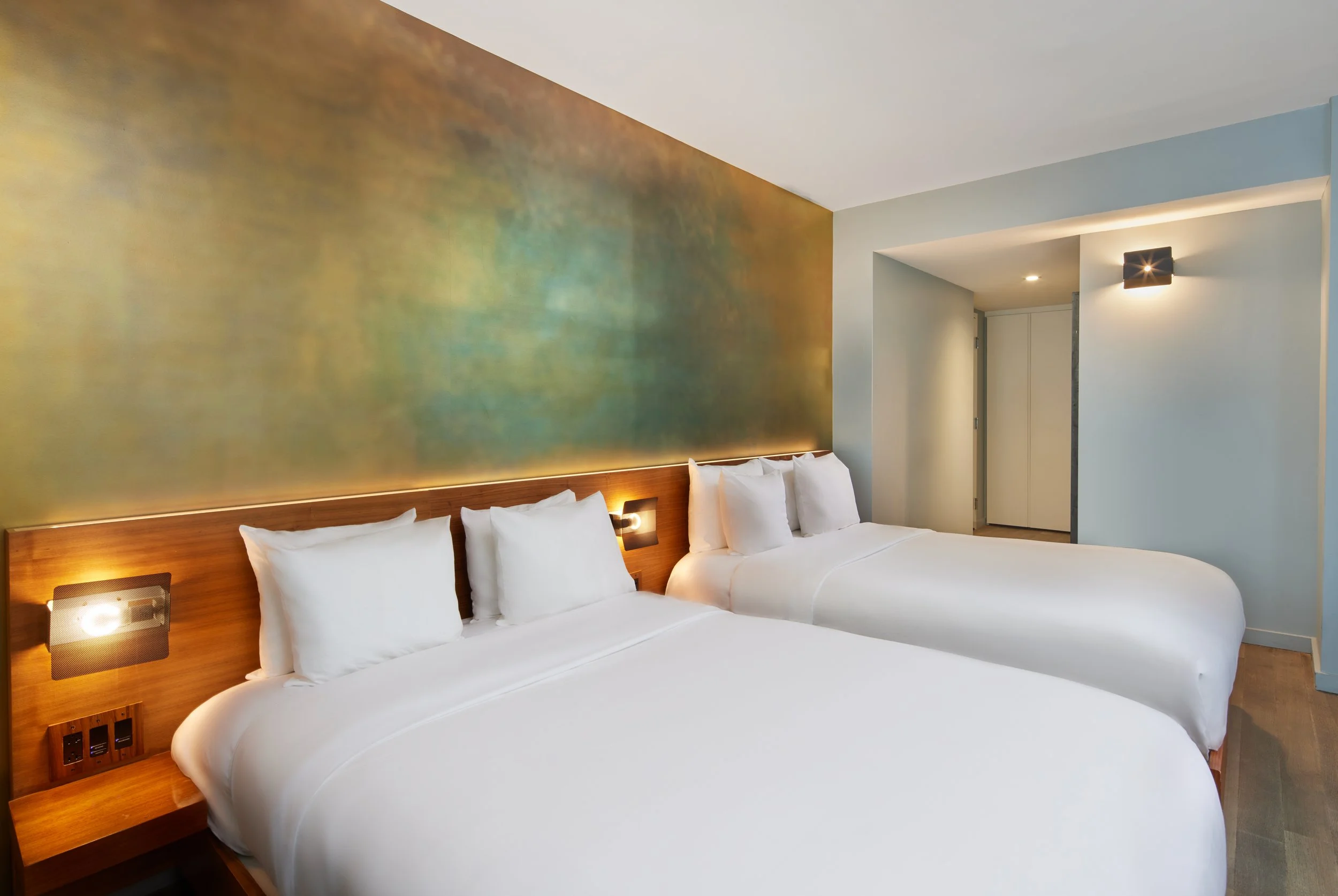 Two neatly made white beds with wooden headboards in a modern hotel room with abstract greenish-golden wall art and ambient lighting.