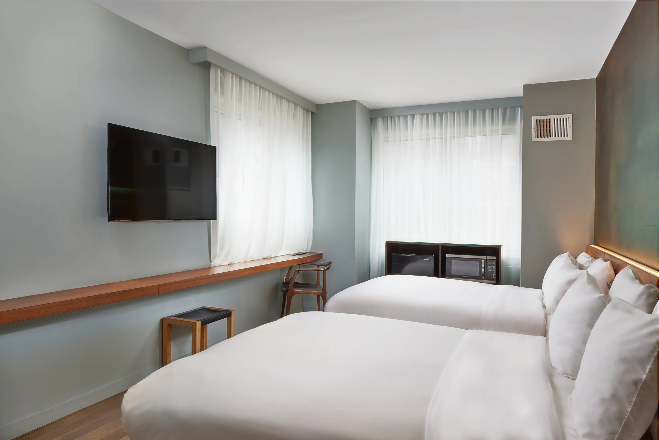Hotel room with two white beds, a wall-mounted TV, a window with sheer curtains, a small wooden desk and chair, and a microwave and mini refrigerator in the corner.