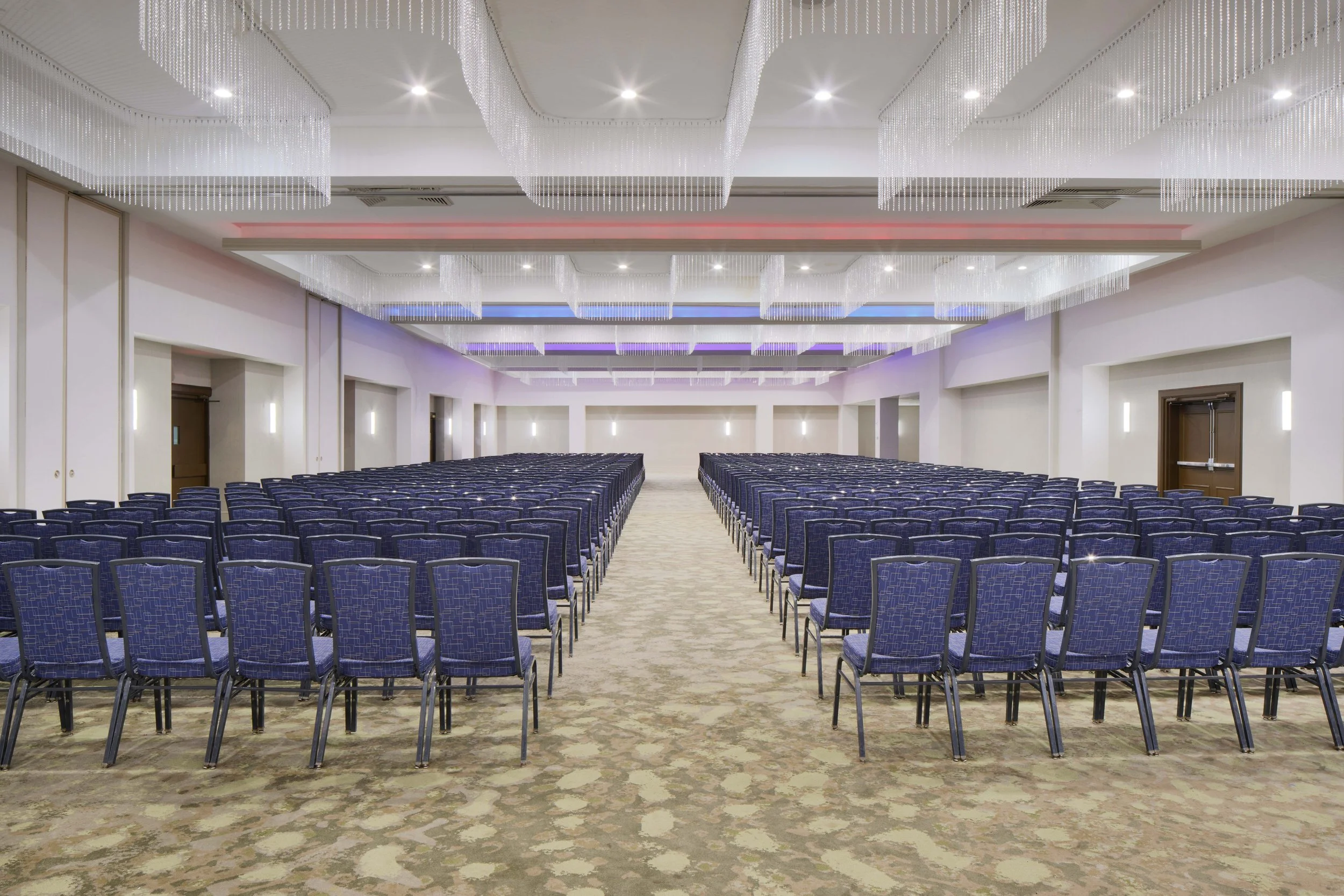 An empty conference or event hall with rows of blue chairs facing a stage or presentation area, decorated with modern ceiling lights and colorful ambient lighting.