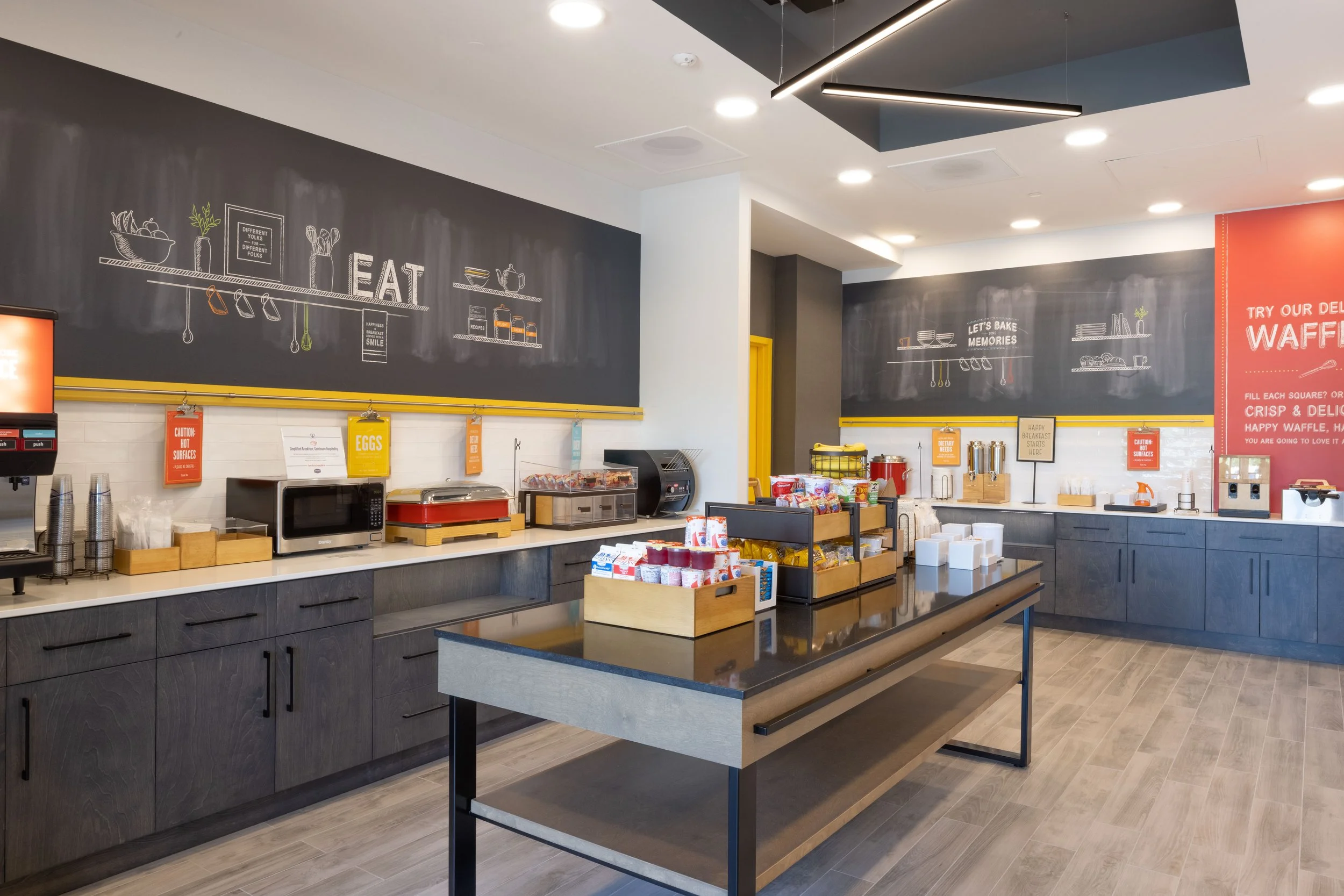 A modern breakfast buffet area with dark cabinets, a white countertop, and chalkboard wall art. The area has various breakfast items, a microwave, and signs indicating different sections for eggs, waffles, and drinks.