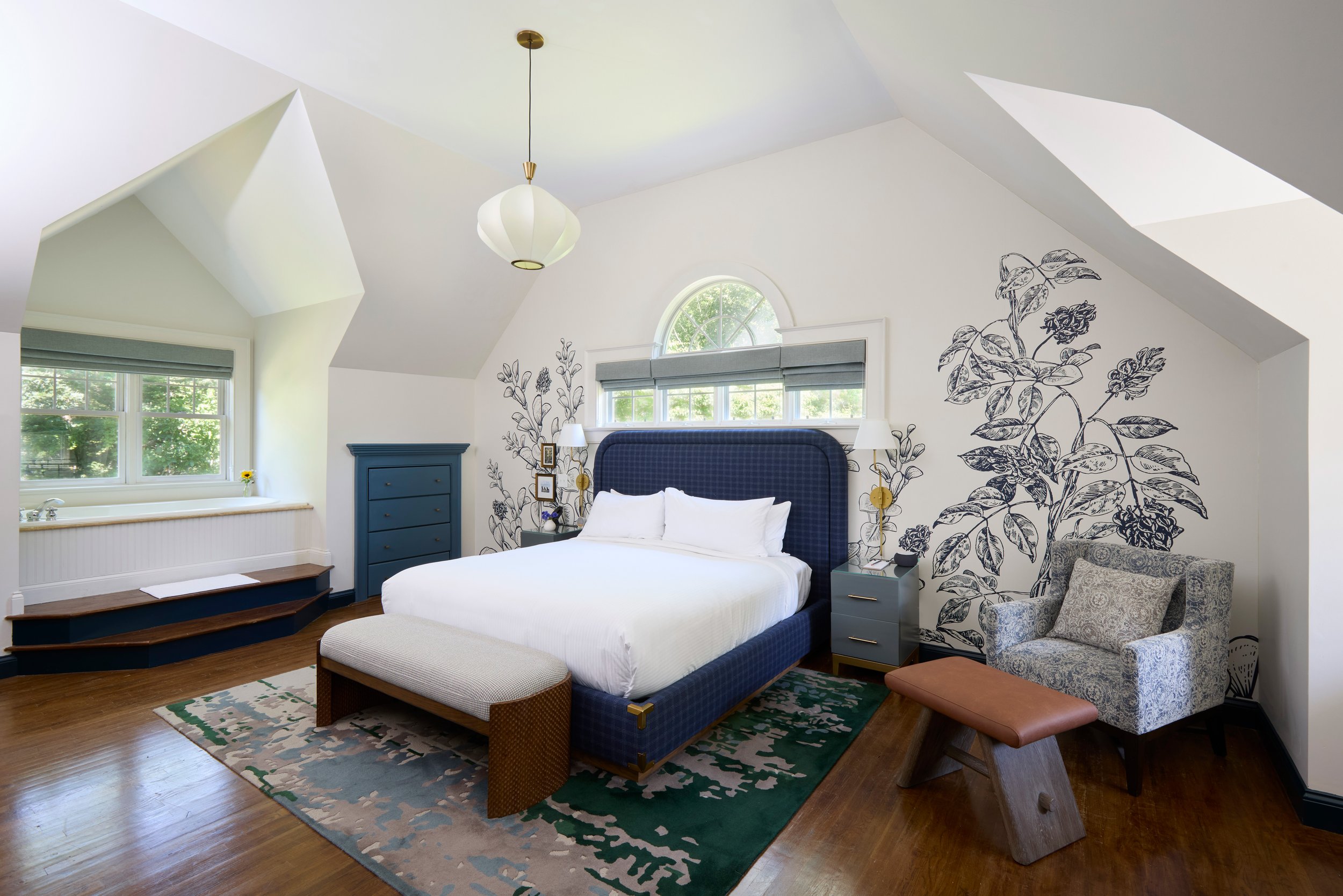 Bedroom with white walls, vaulted ceiling, and hardwood floors. Features a blue upholstered bed with white bedding, a gray bench at the foot of the bed, and a reading chair with a brown ottoman. Two nightstands with lamps and decorative wall art of b