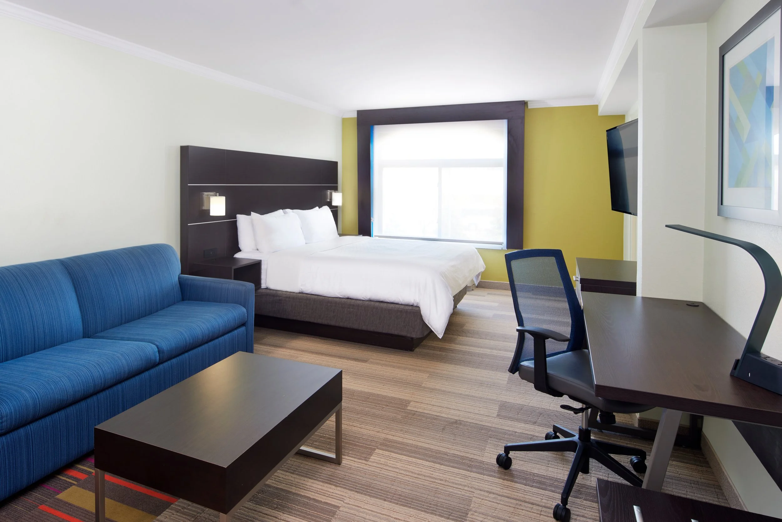 Hotel room with a large bed, blue sofa, desk with chair, wall-mounted TV, and window with blinds