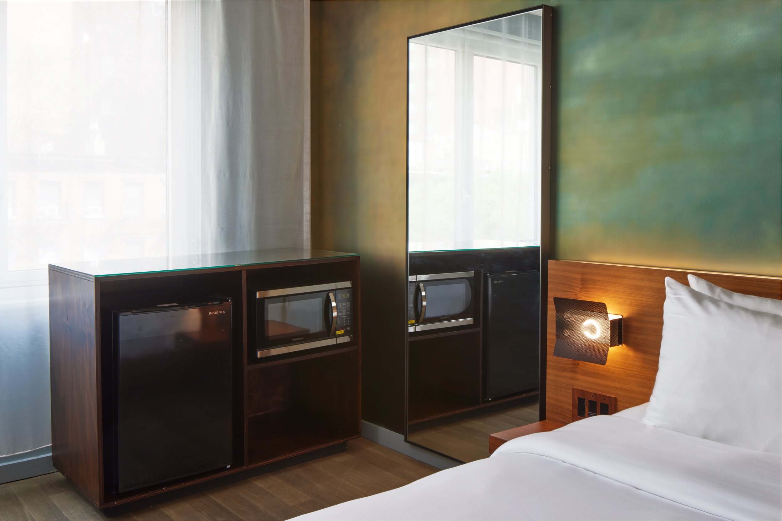 hotel room with a bed, a wall-mounted lamp, a large mirror, a microwave, a mini fridge, and a glass-topped cabinet near a window with sheer curtains.