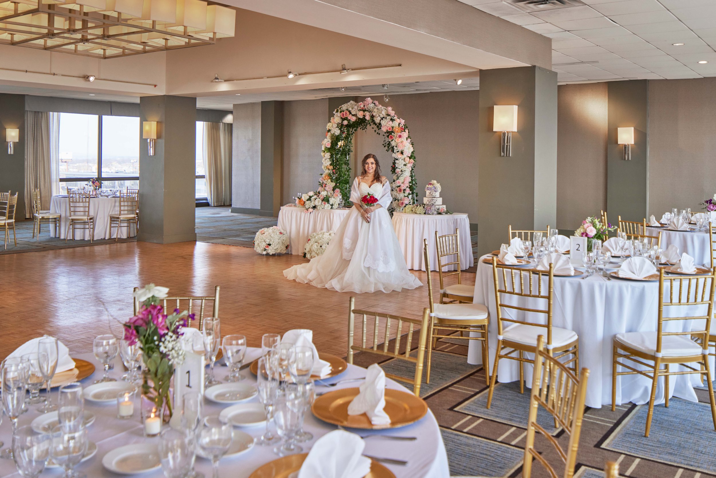 A bride in a white wedding gown holding a bouquet, standing in a decorated reception hall with round tables, gold chairs, and floral centerpieces.