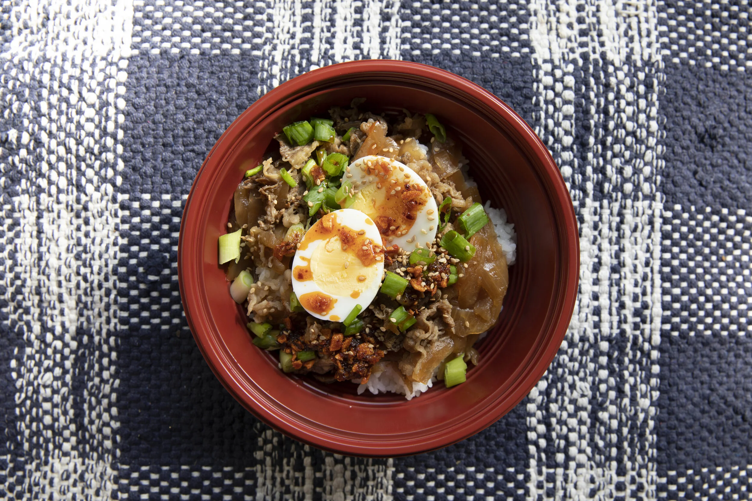 A bowl of Asian-inspired rice topped with sliced boiled eggs, chopped green onions, seasonings, and cooked meat on a black and white checkered cloth