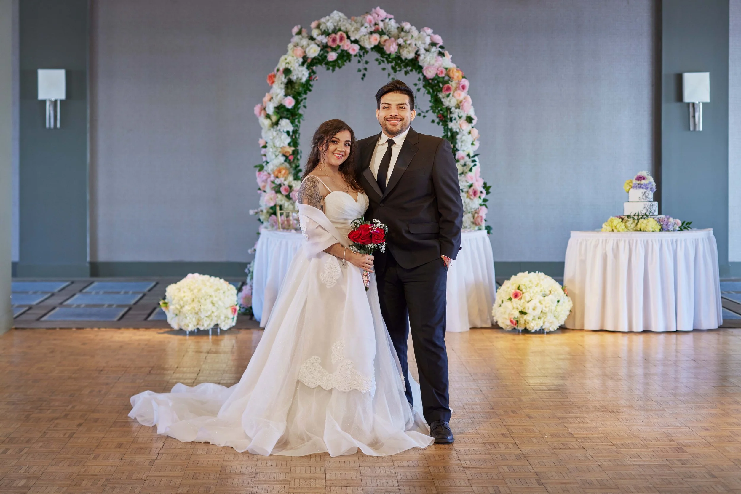 A newlywed couple smiling for a photo in a decorated wedding venue. The bride wears a white wedding gown and holds a bouquet of red roses. The groom wears a black suit and tie. Behind them is a floral arch with pink and white flowers, and there are t