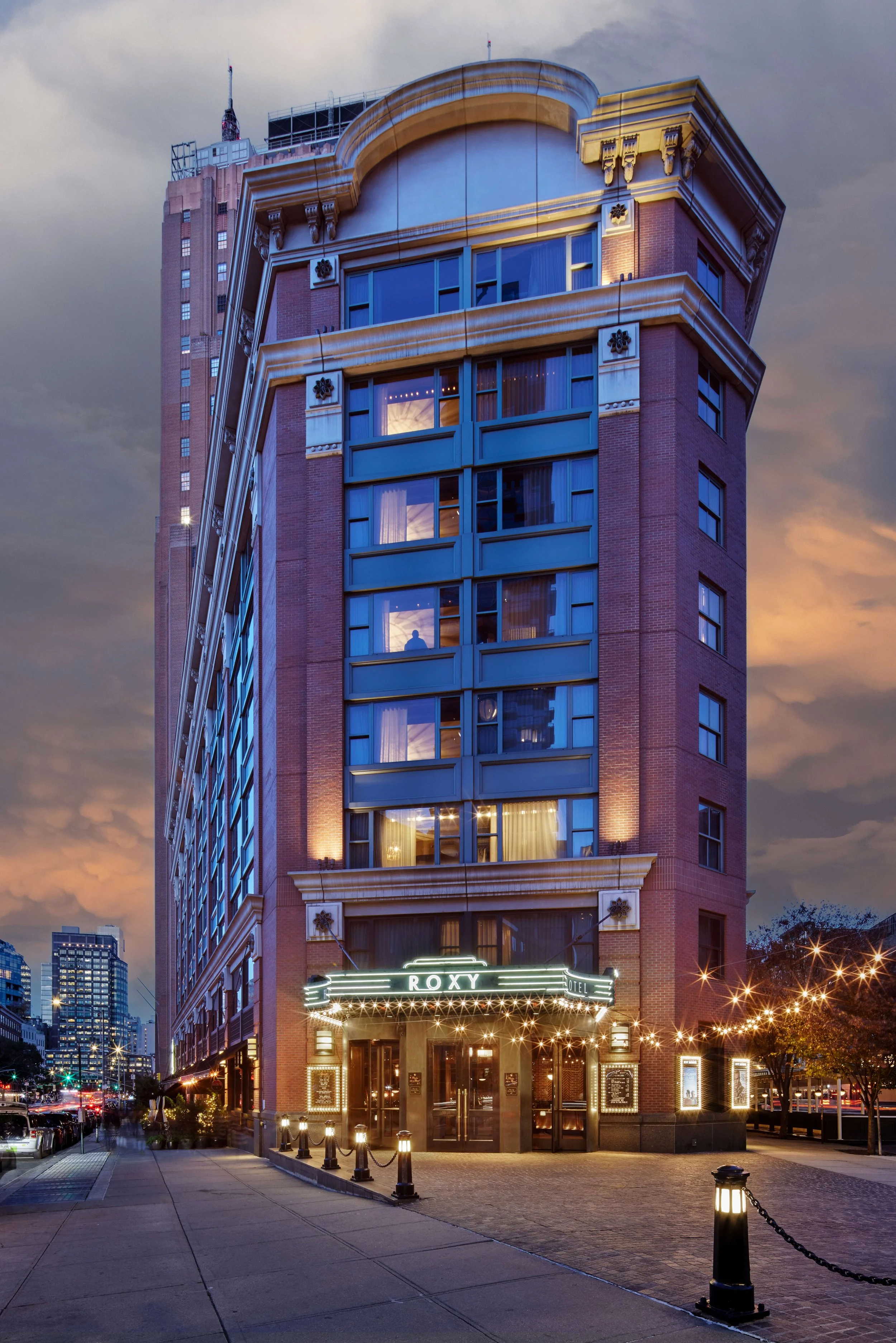 A tall, multi-story hotel building with illuminated signage reading 'ROXY' at the entrance, during dusk. The building features brick and modern glass windows, with decorative architectural details. Streetlights and trees line the sidewalk in front.