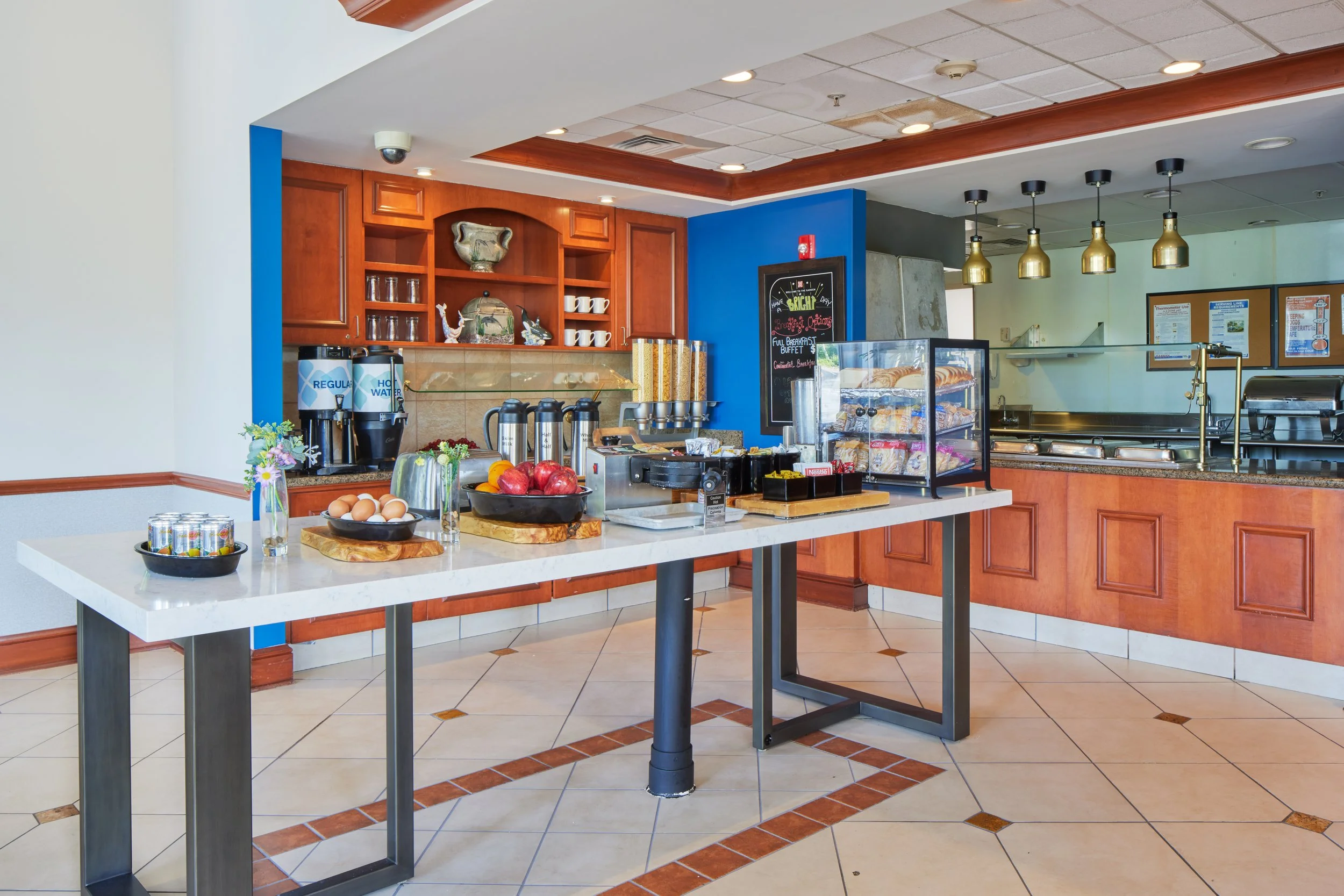Breakfast buffet area with apples, eggs, cereal, bread, and coffee, in a dining room with tile floors and wooden accents.