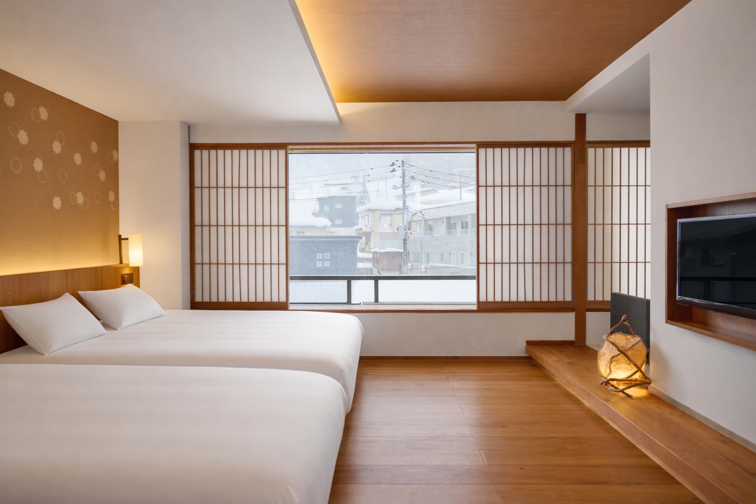 A modern hotel room with a large bed, white bedding, and pillows, illuminated by a wall-mounted light. It features a large window with Japanese-style shoji screens showing a snowy urban scene outside. The room has wood flooring, a mounted TV, and a d