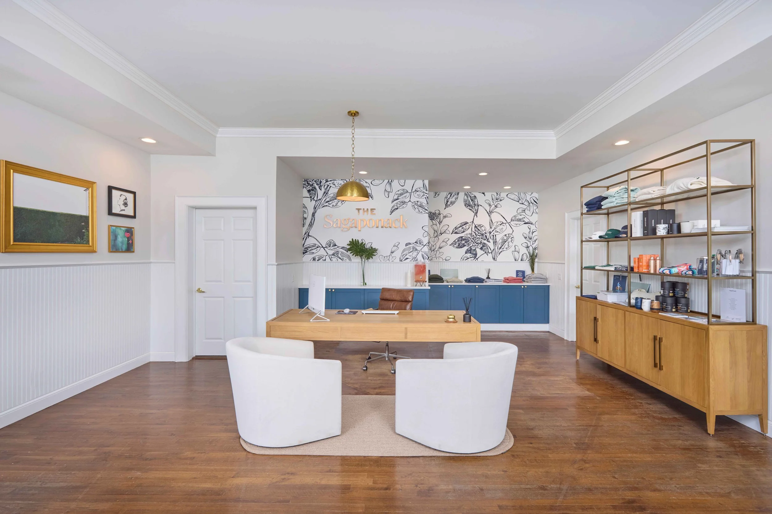 Office reception area with a wooden desk, two white chairs, a blue cabinet, and a large shelving unit with folded towels and products. The wall behind has a sign: 'The Sagaponack', and there is black and white botanical wallpaper.