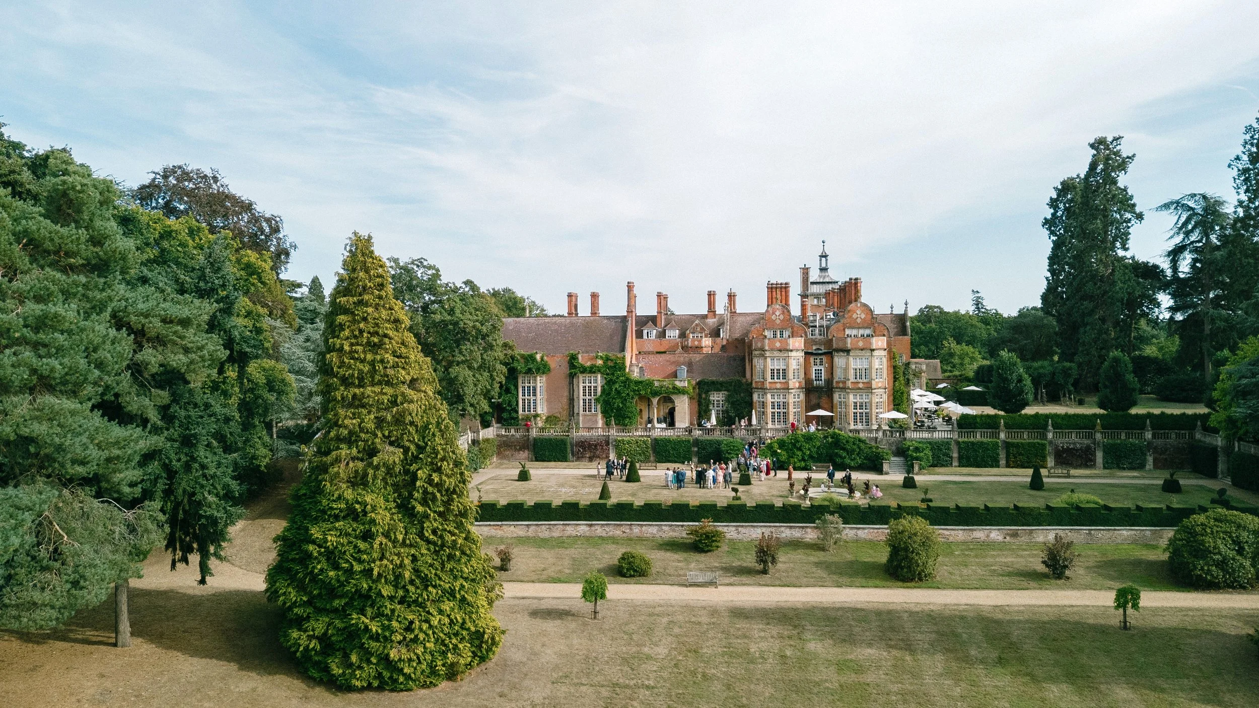 A large historic mansion surrounded by greenery, including various tall trees, with a well-maintained lawn and a group of people gathered in front of the building on a sunny day. At Tylney Hall wedding photography