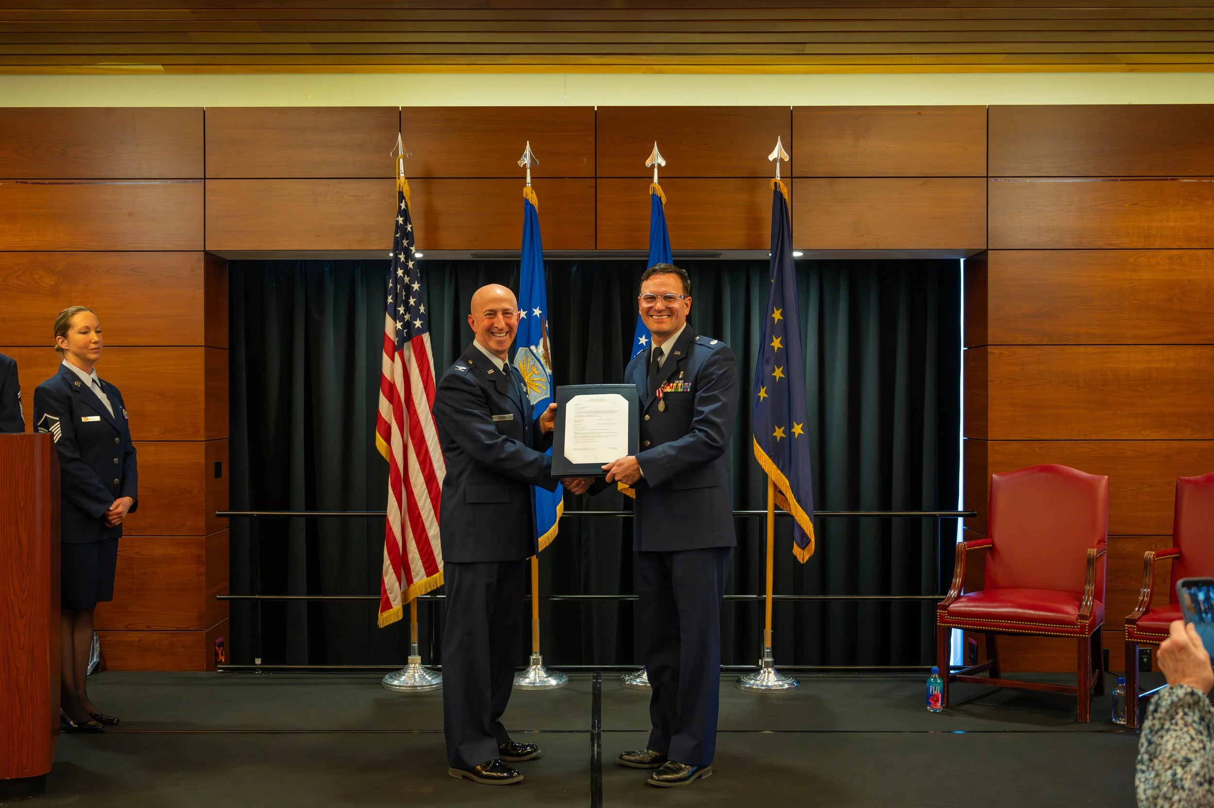 Two uniformed men smiling and shaking hands during a formal ceremony, with flags and curtains behind them, and women in uniform standing to the sides.