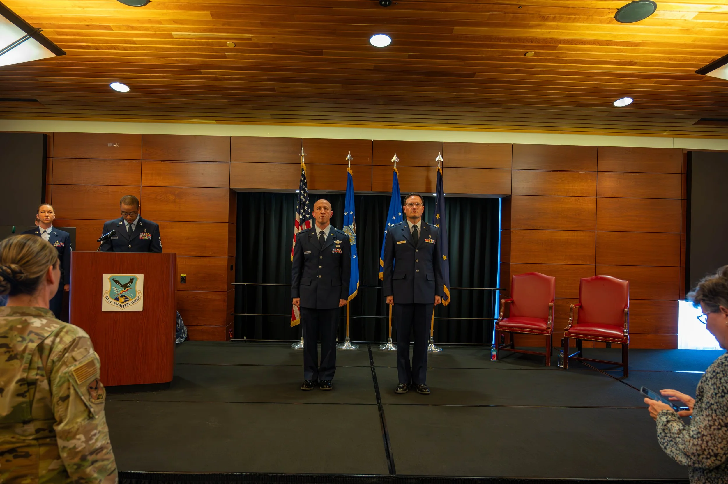Four military personnel in uniform standing on a stage in front of flags, with audience members watching.