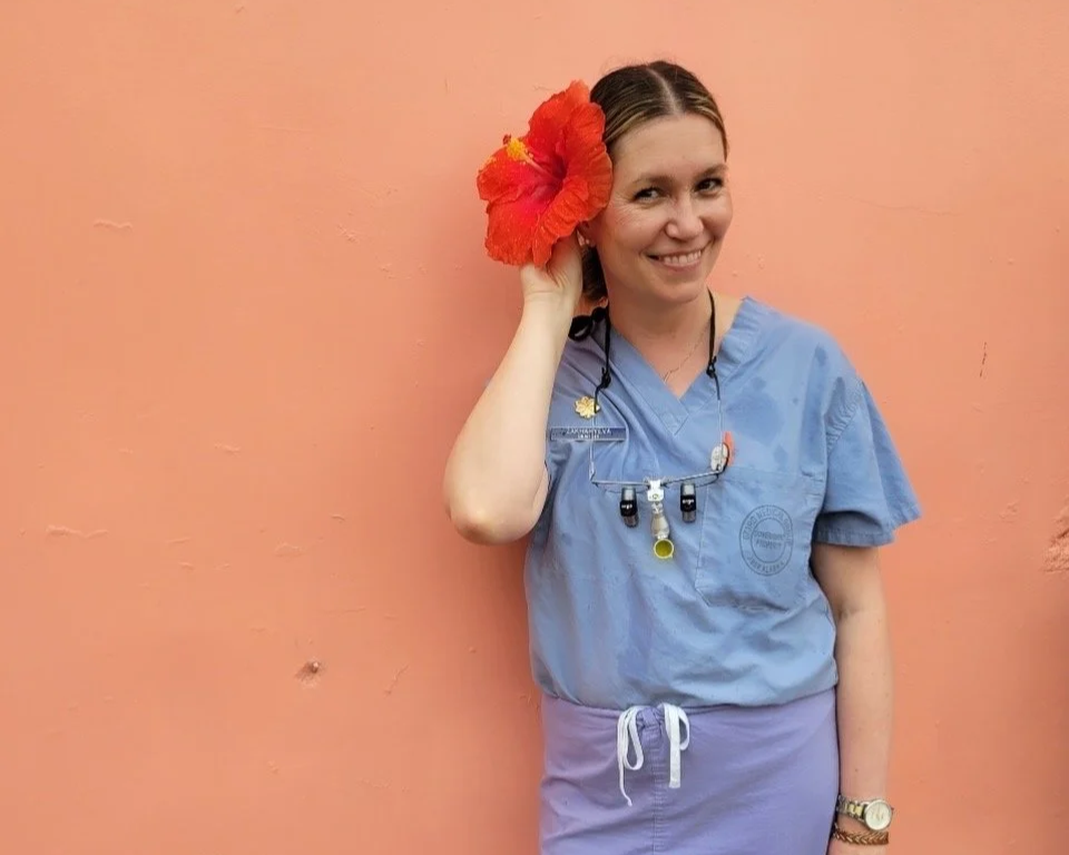 A woman in medical scrubs with a large red hibiscus flower in her hair, smiling, standing against a pink wall.