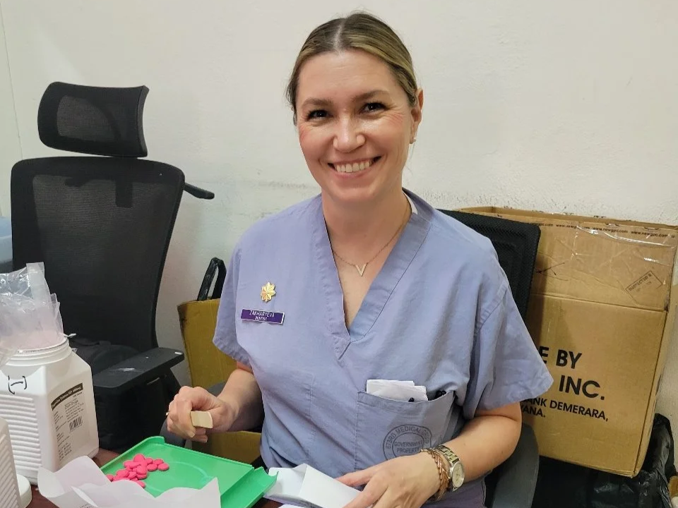 A smiling woman in purple scrubs sitting at a desk with medical supplies, a black office chair, and a cardboard box behind her.