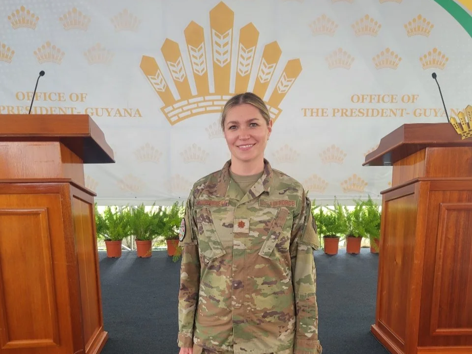 A woman in military uniform standing in front of a backdrop with gold crown symbols and the text 'Office of the President Guyana' and 'Office of the President - Guyana'. She is smiling and positioned between two wooden podiums.