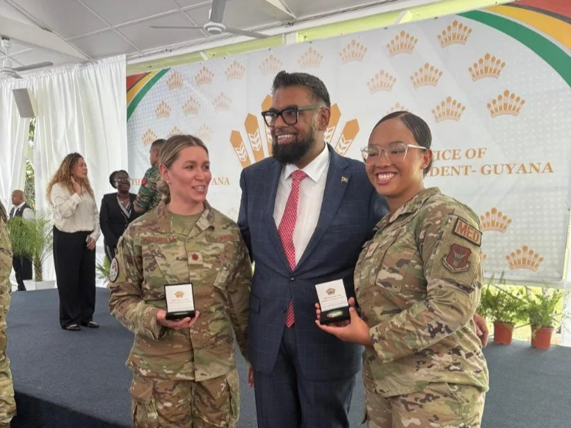 Two female soldiers in camouflage uniforms holding medals, standing next to a man in a suit with glasses, during an award ceremony in Guyana.