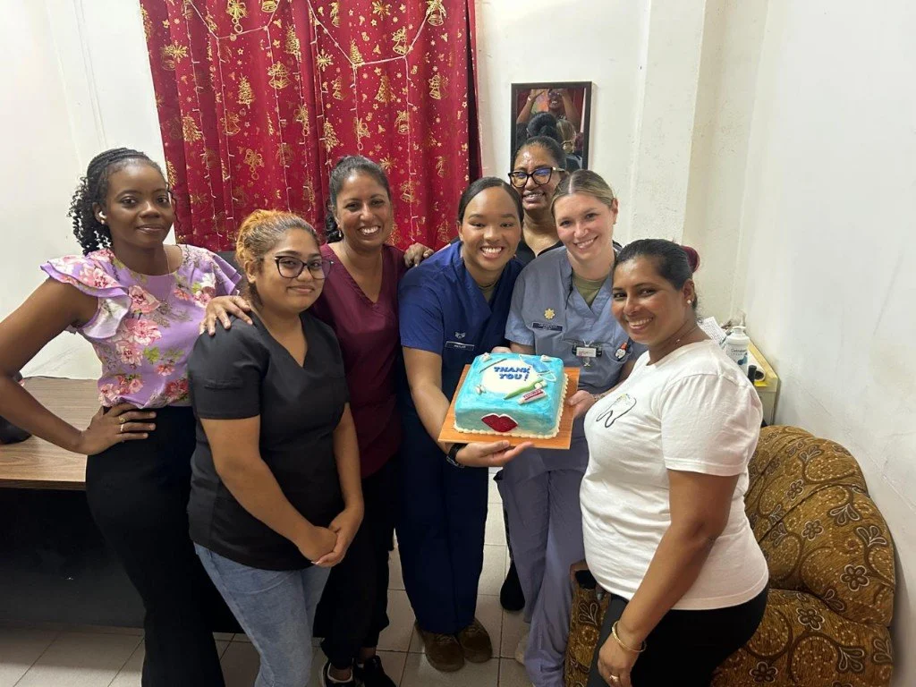 Group of seven women celebrating with a cake that says "Thank You!" in a room with red curtains and a mirror.