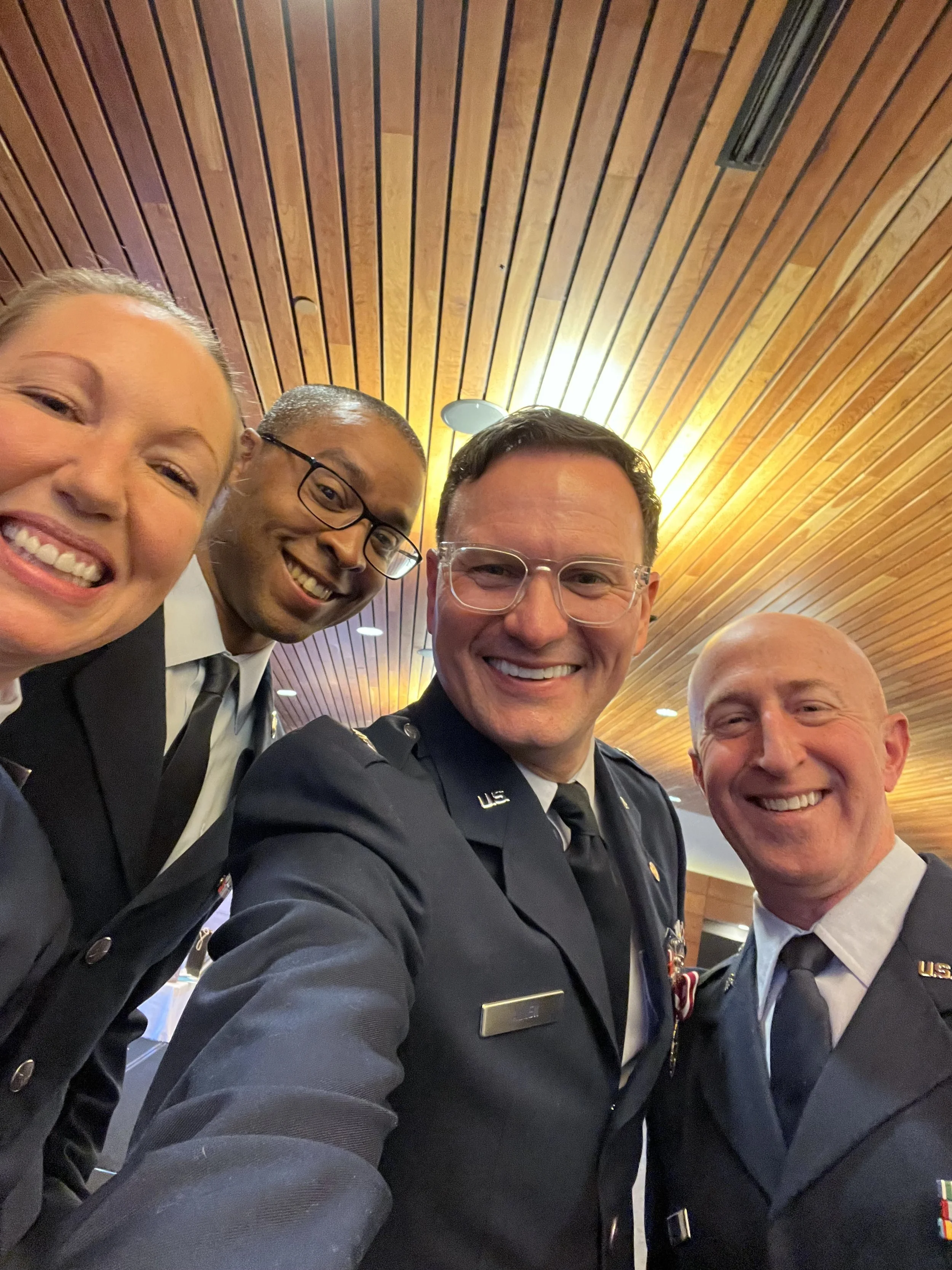 A group of four smiling people, including two men in military uniforms, a woman, and another man, taking a selfie together indoors with a wooden ceiling in the background.