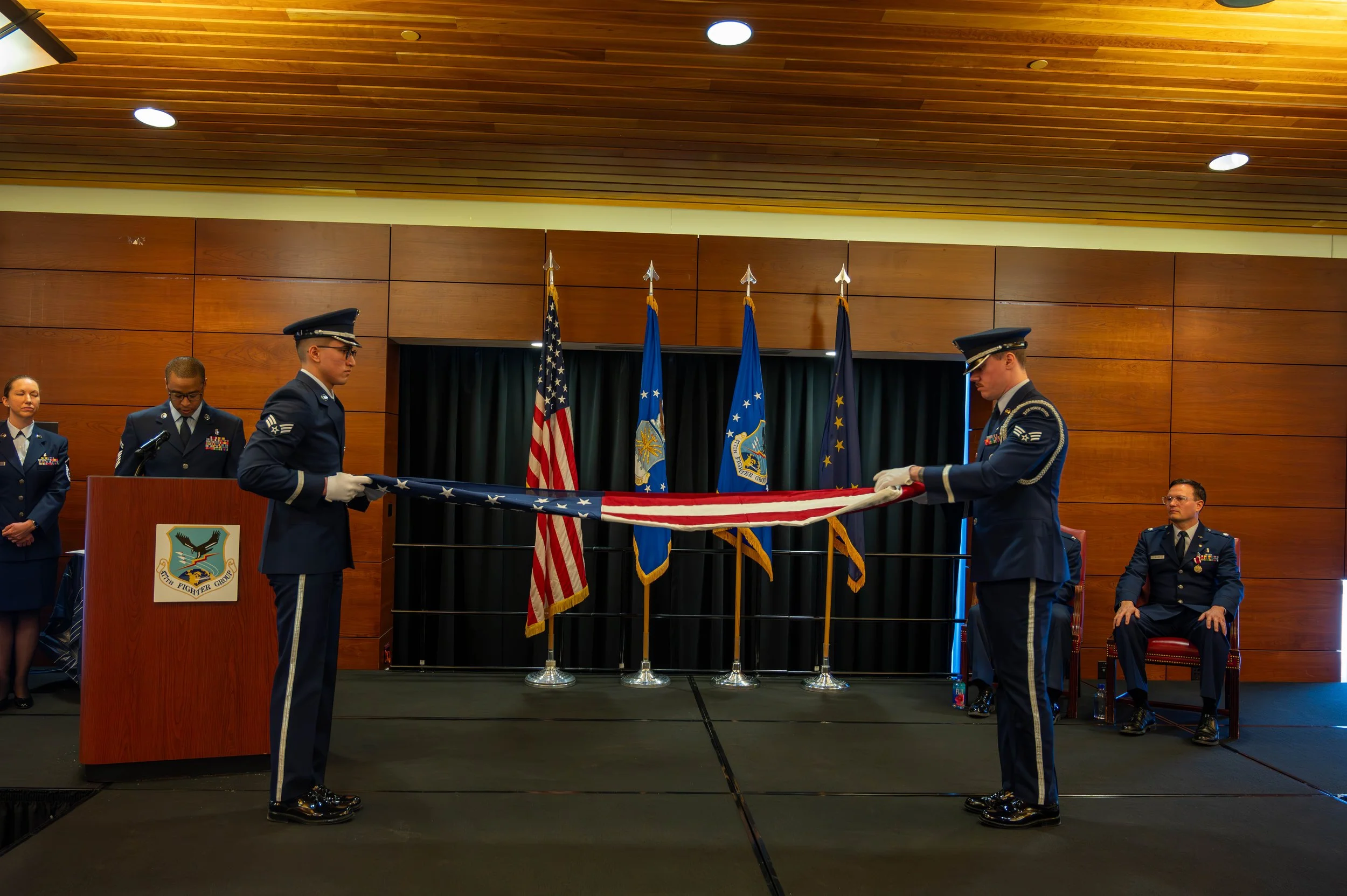 Military personnel in uniform participating in a flag ceremony with American and military flags on a stage with wooden walls and a curtain.