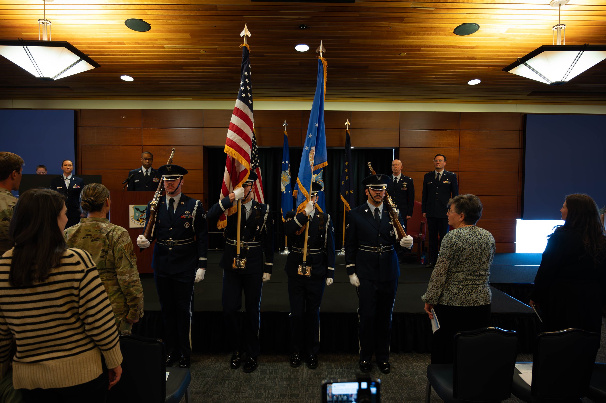 Military ceremony with uniformed personnel carrying flags, audience members watching, and a stage backdrop with blue and wooden panels.