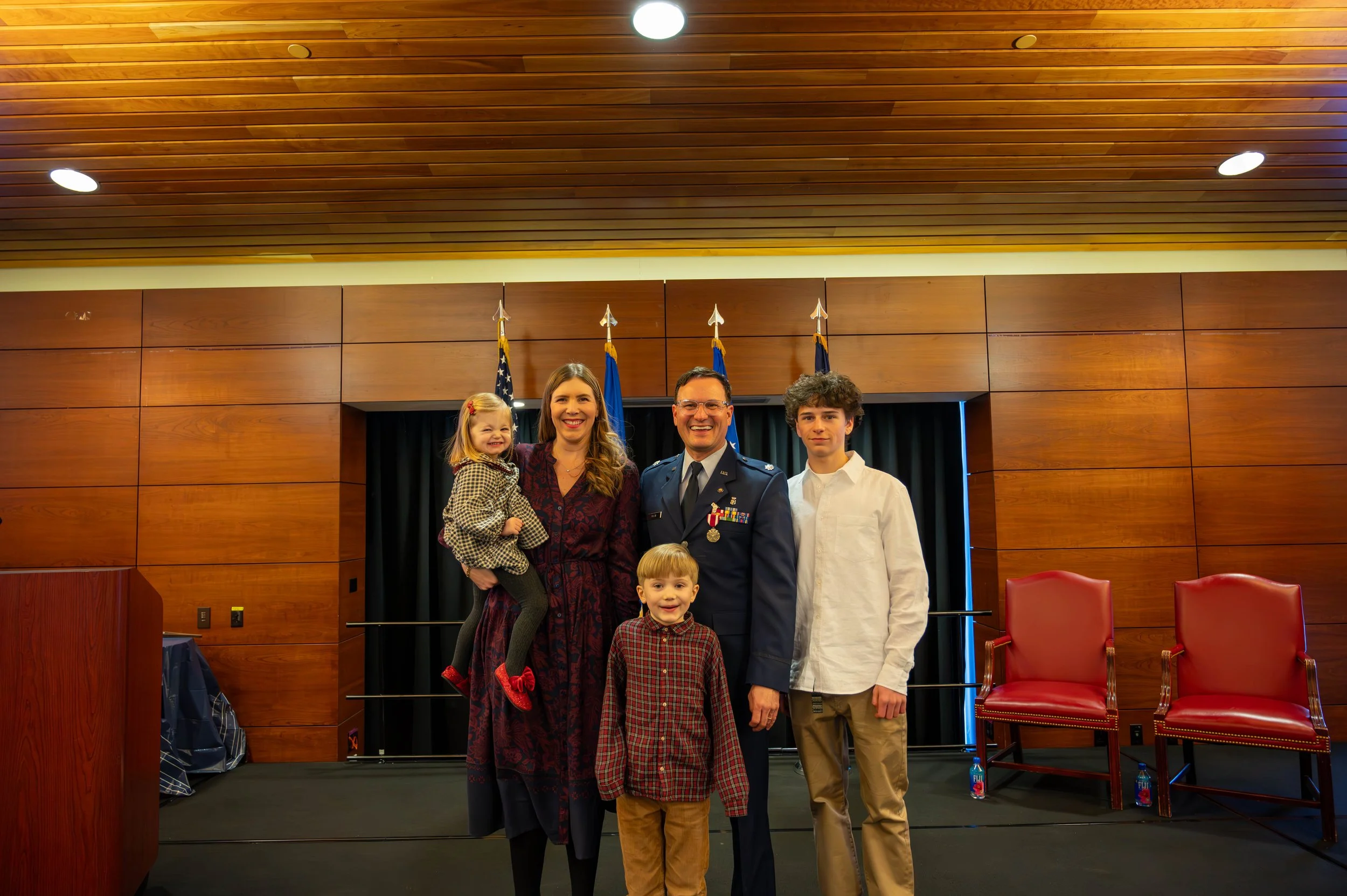 A group of five people, including a man in military uniform and four children, standing on a stage in a wooden-paneled room with flags behind them.