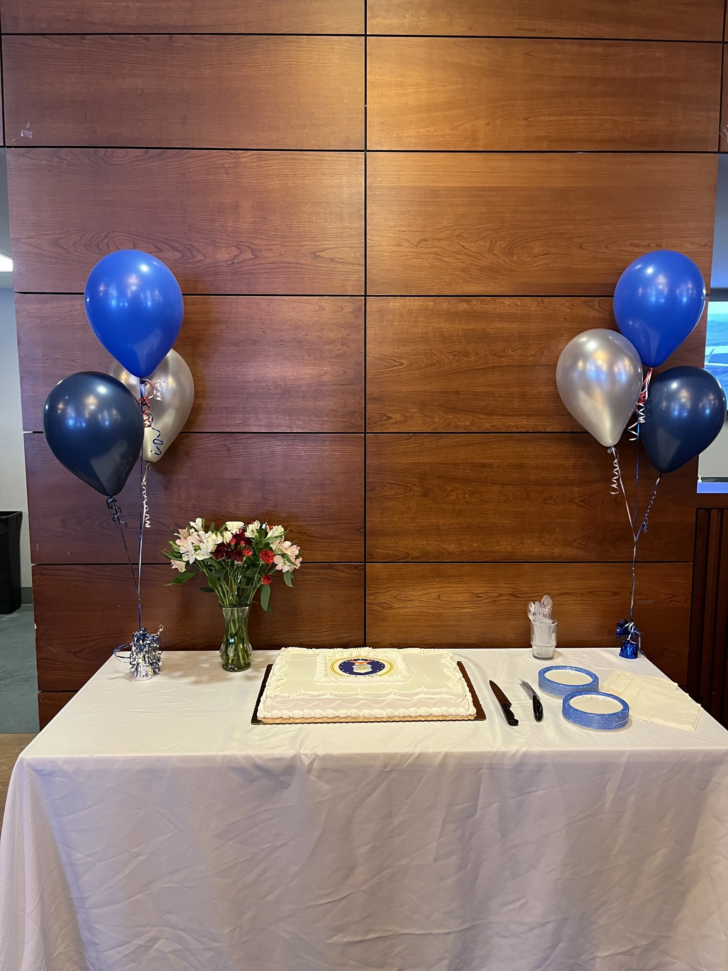 A table with white tablecloth, decorated with a cake, flowers, and party supplies, against a wooden wall with balloons on either side.