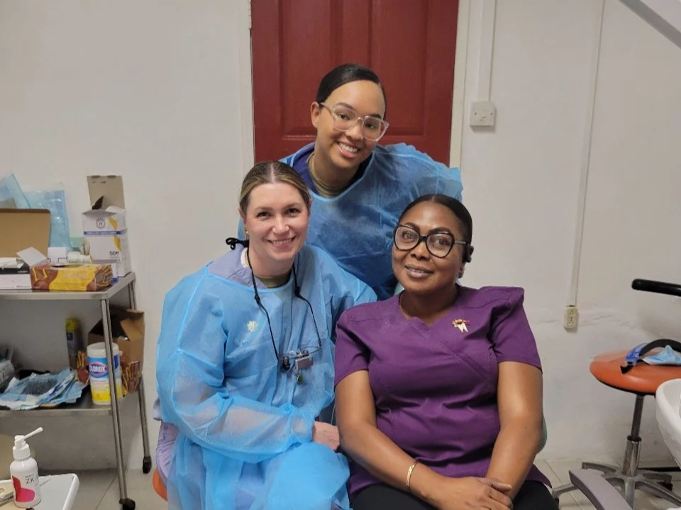 Three women, one seated and two standing, in a medical setting. The seated woman is wearing glasses and a purple medical uniform, and the two standing women are dressed in blue medical protective gear, smiling at the camera.