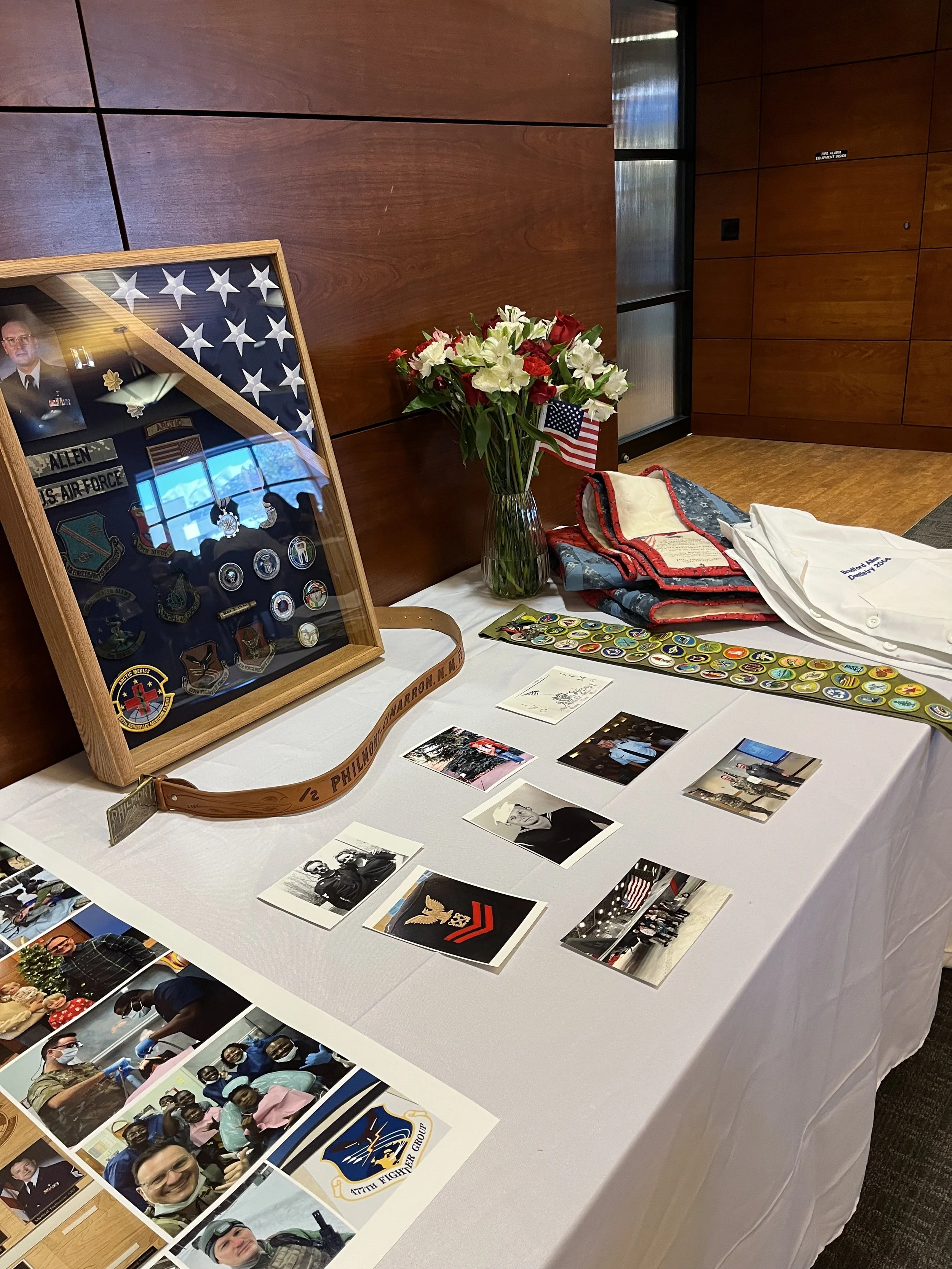 A table with a display of photographs, military patches, and memorabilia, including a framed display of military patches and medals, a bouquet of red, white, and green flowers with small American flags, and various patches and pictures related to mil