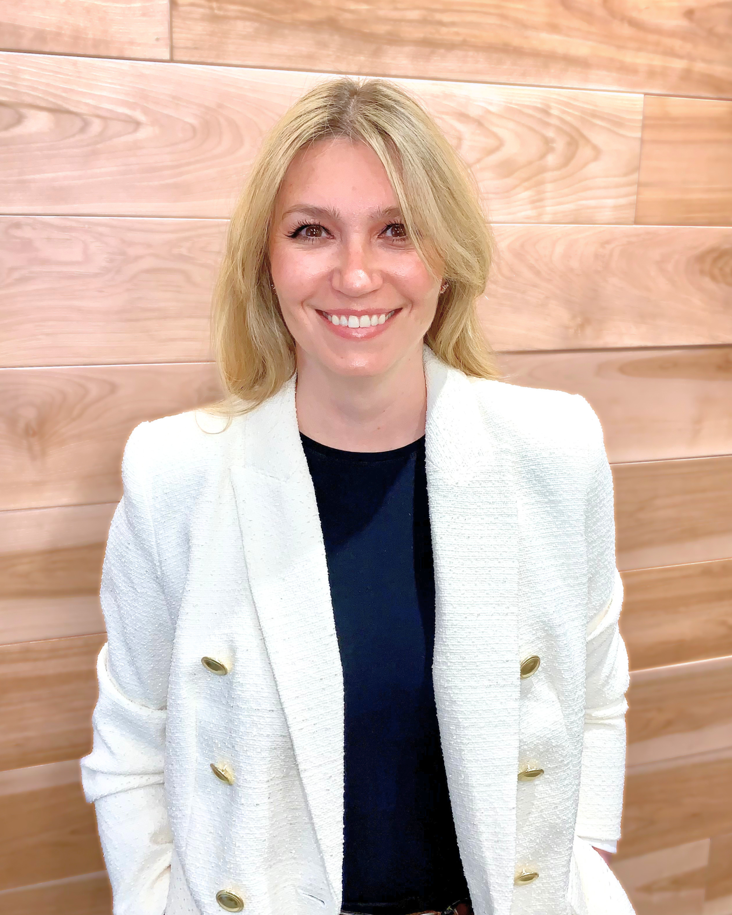 A woman with blonde hair, smiling, wearing a white textured blazer with gold buttons, standing in front of a wooden wall.