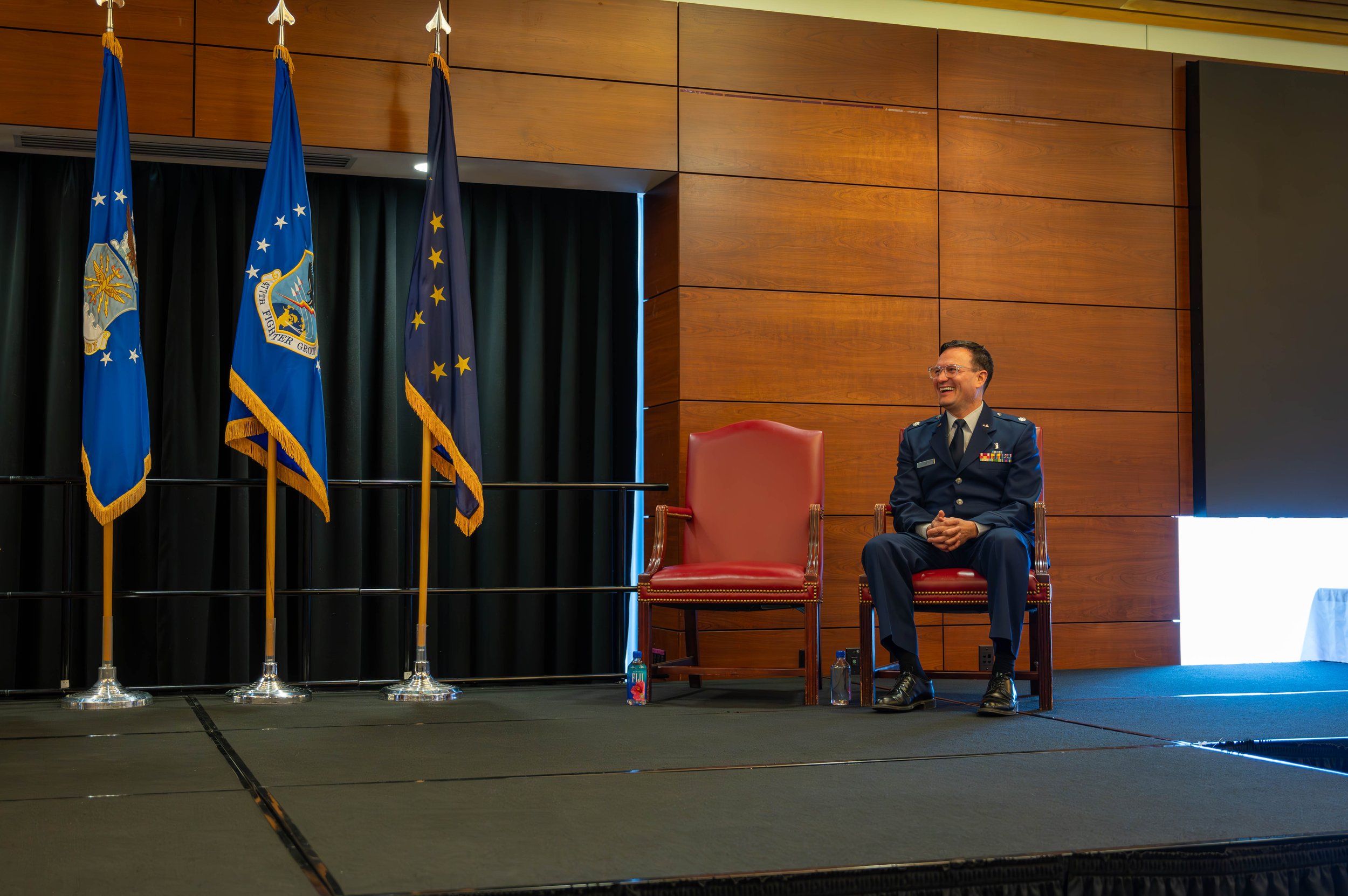 A man in a military uniform sitting on a chair on a stage, smiling. Behind him are three flags, two blue and one dark blue, with golden fringes. The stage has black curtains and wooden paneling.