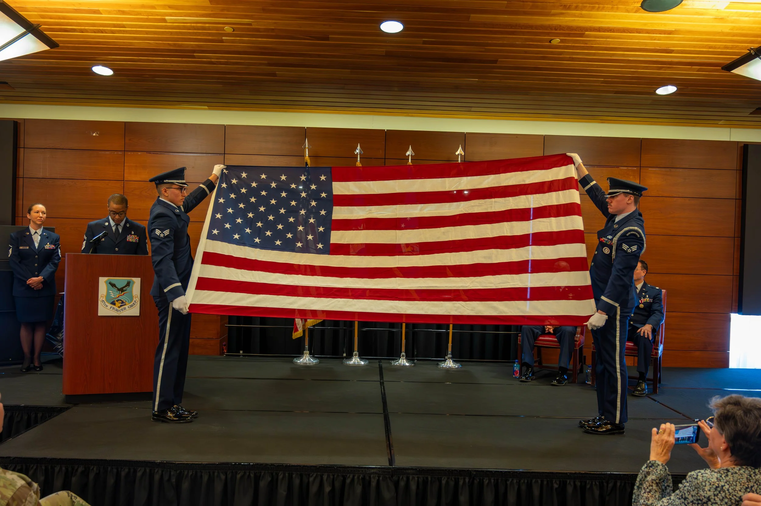 Military officers in uniform folding a large American flag during a ceremony in a room with wooden walls and a wooden ceiling. Several attendees are present, some taking photos.