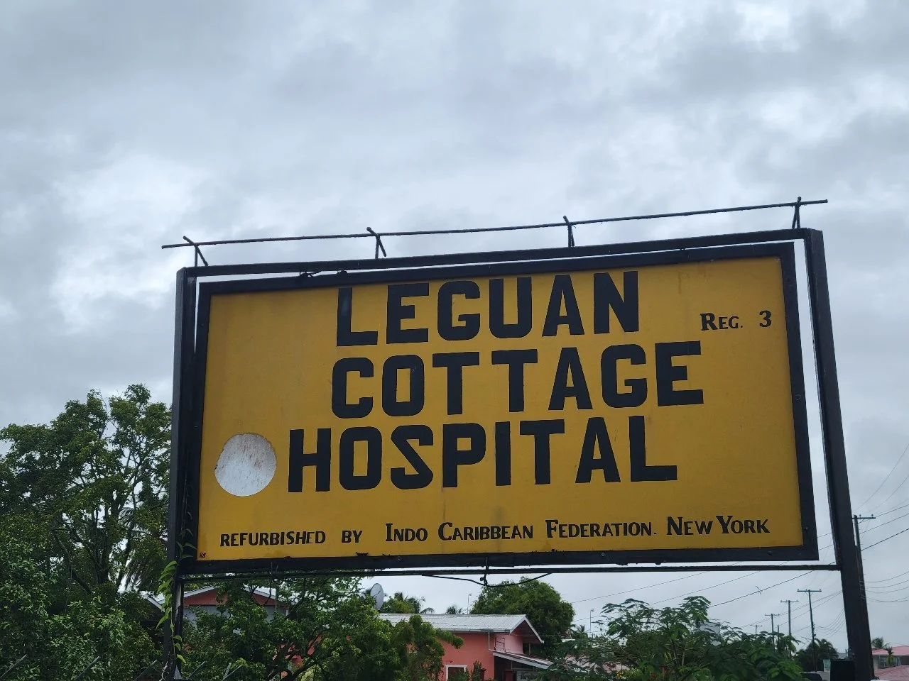 Yellow signboard reading 'Leguan Cottage Hospital' with additional text indicating refurbishment and registration details, against a cloudy sky and nearby trees and buildings.