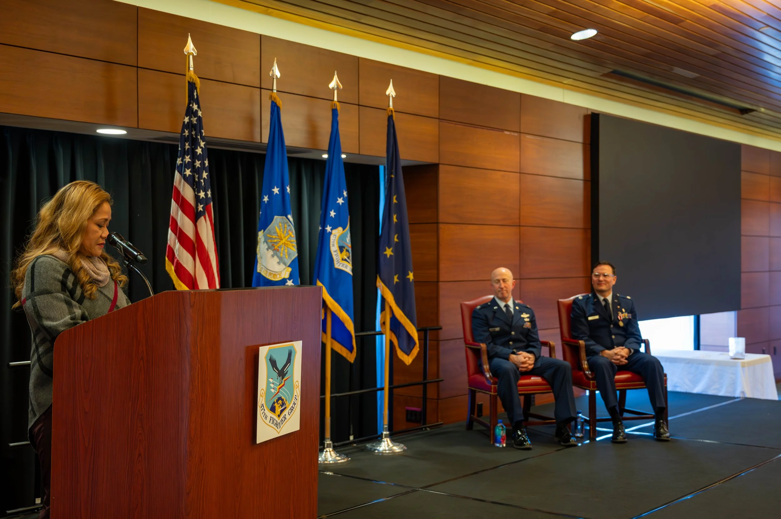 A woman stands at a wooden podium with a military crest, reading or speaking into a microphone while a panel of three men in military uniforms, two seated and one standing, listen. Behind them are four flags, including the American flag and others wi