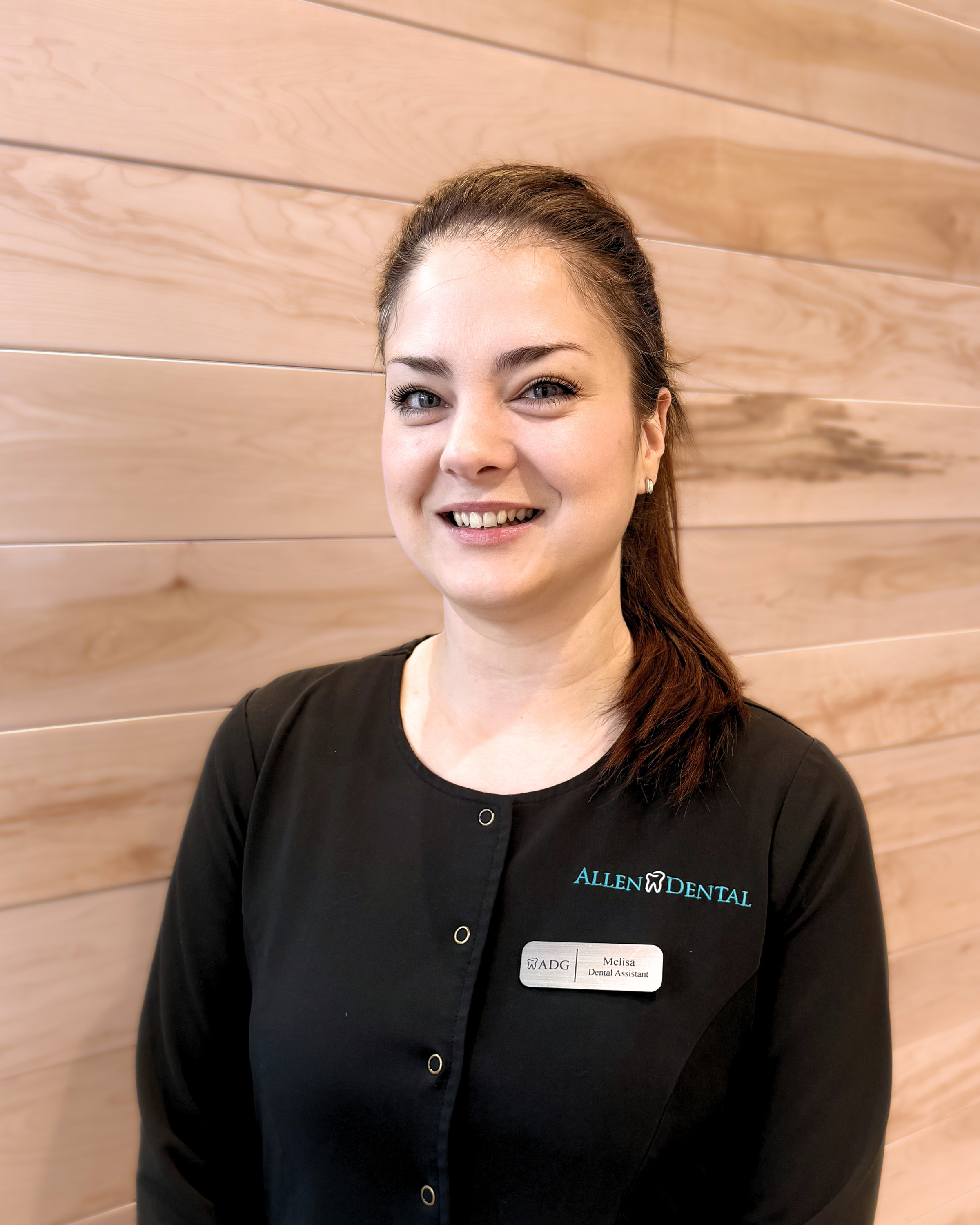 A woman smiling, with brown hair in a ponytail, wearing a black dental assistant uniform with a name tag that reads Melisa. The uniform has the logo "ALLEN DENTAL" on the upper left side. She is standing in front of a wooden wall.