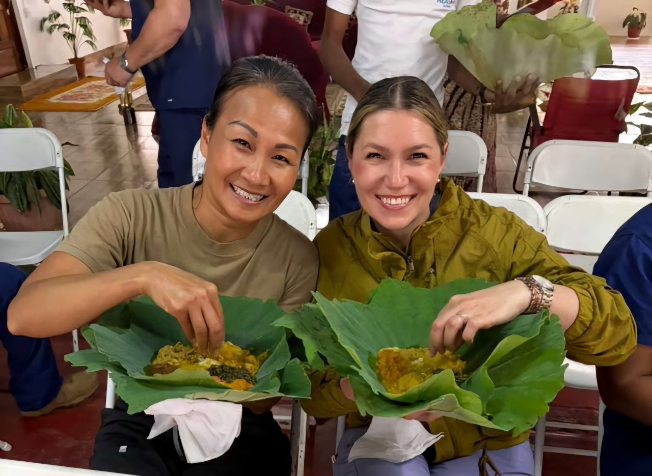 Two women smiling and holding food dishes wrapped in banana leaves at a gathering, with other people in the background.