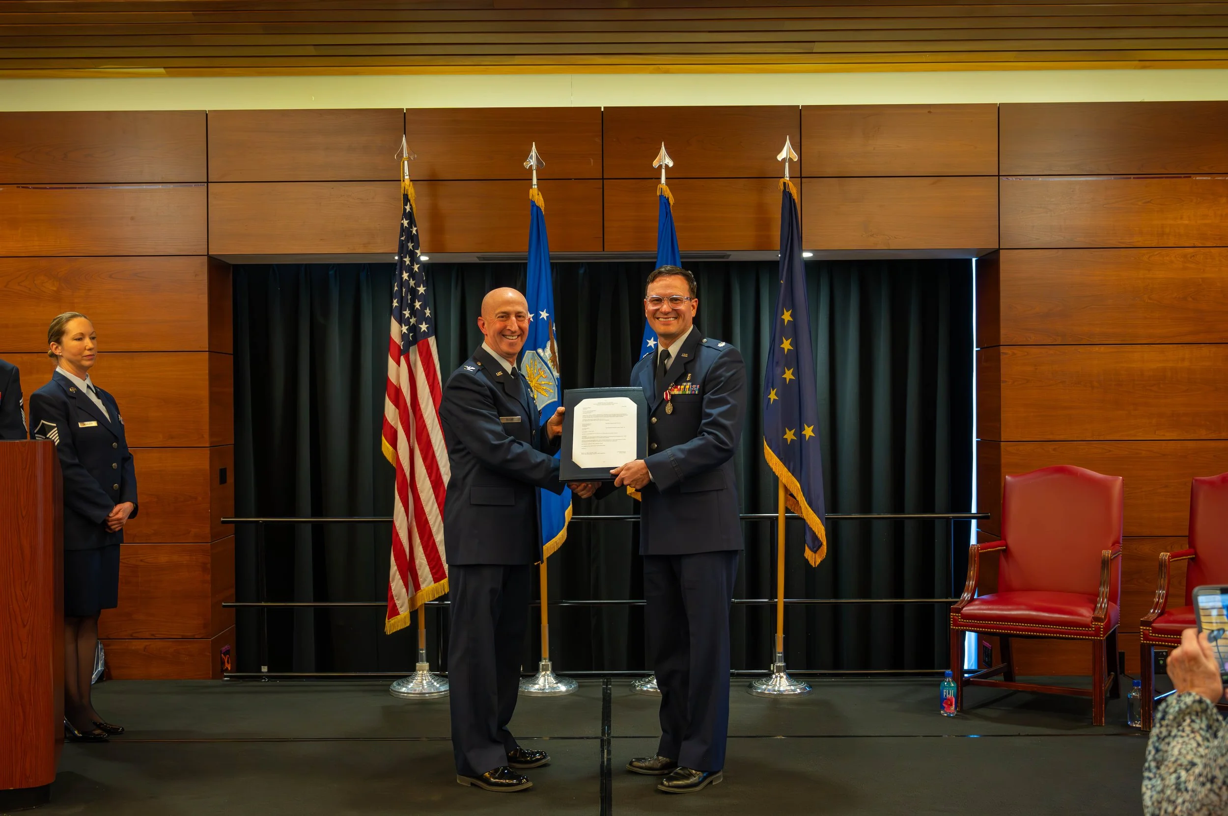 Two men in military uniforms are smiling and shaking hands during a formal ceremony, with flags in the background. A woman in uniform is standing on the left, and a person taking a photo with a phone is partially visible on the right.