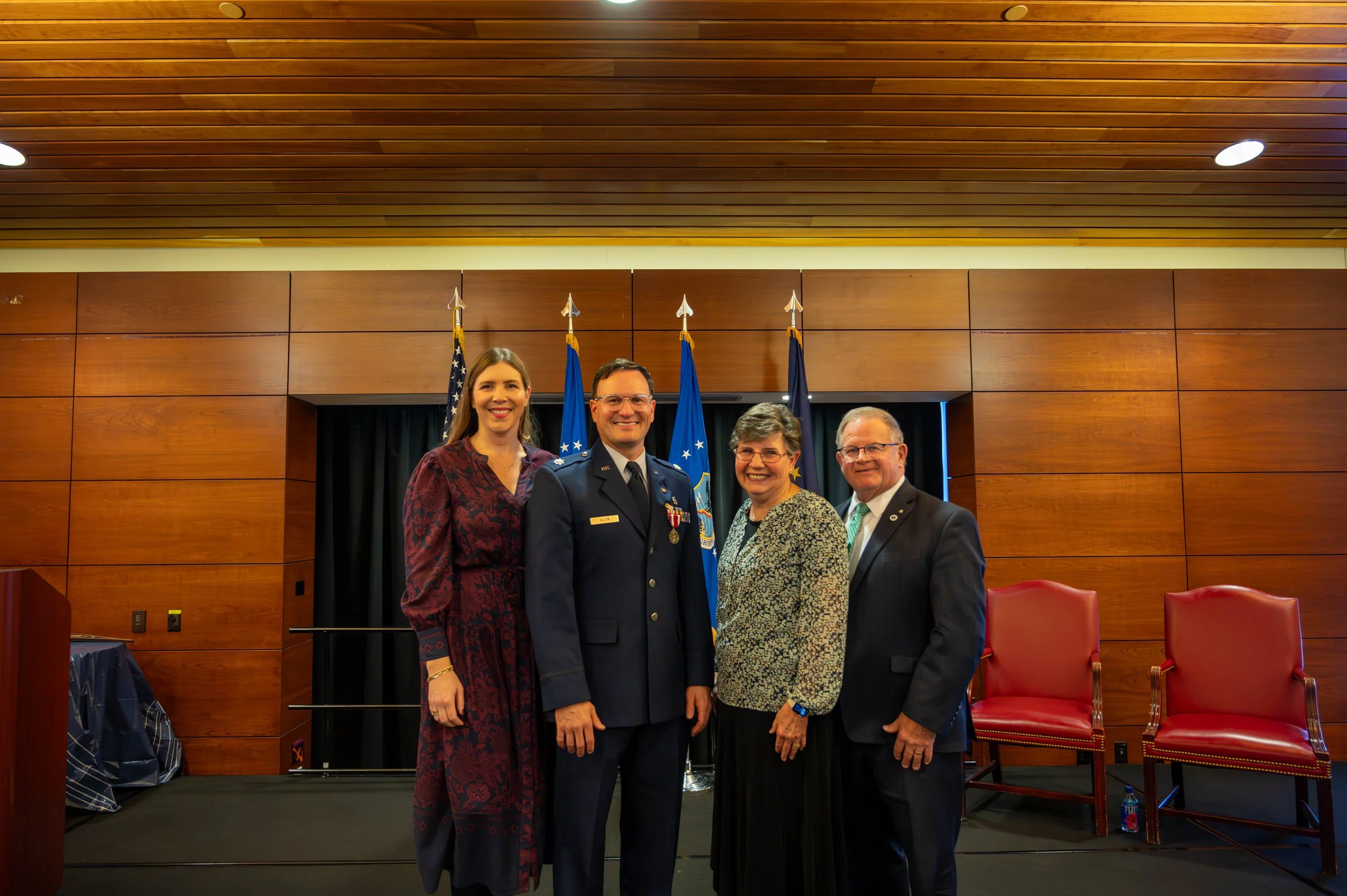 Four people standing together in front of a backdrop with flags, smiling at the camera, in a wood-paneled room with a stage and chairs.