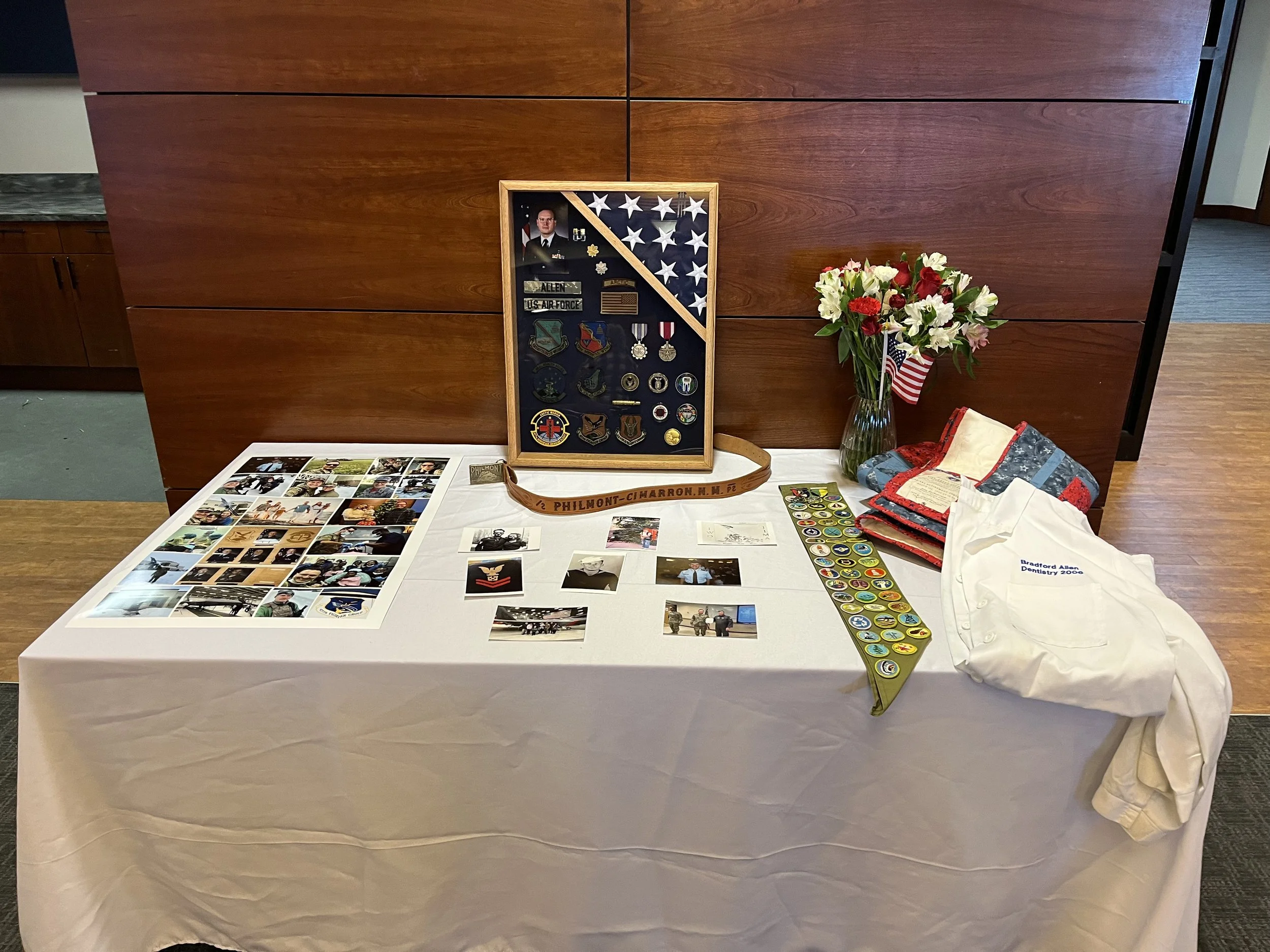 A display table with a collage of photographs, a framed shadow box with military patches and medals, a lanyard, and patches, along with a stack of embroidered fabric and a white cloth bag featuring an American flag pattern, set against a brown wooden
