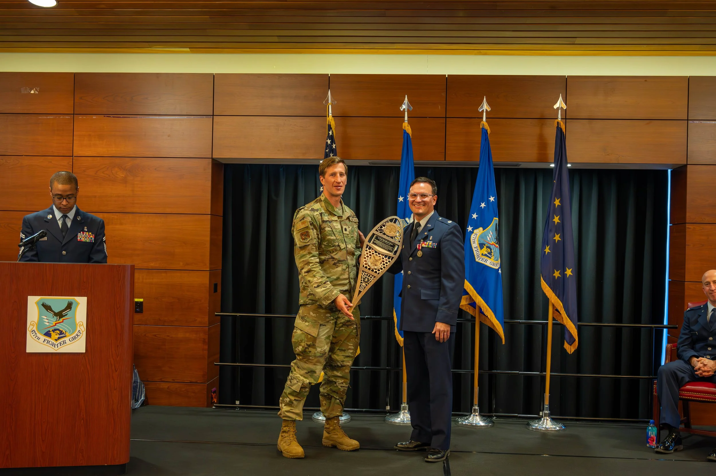 A military award ceremony with two men in uniform, one presenting an award to the other, both smiling, with flags in the background.