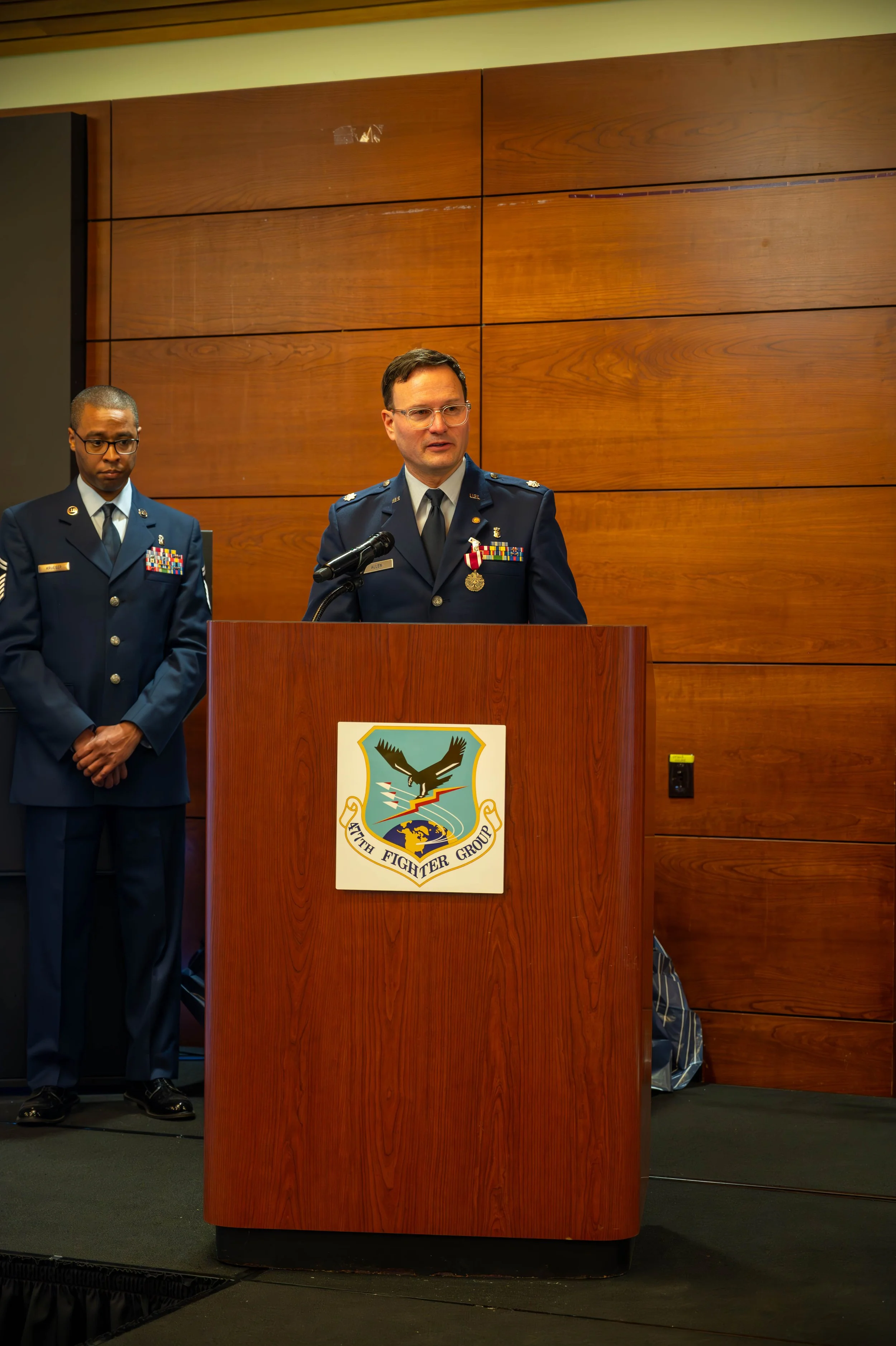 Two men in military uniforms standing behind a wooden podium with the 75th Fighter Group symbol, in a room with wooden panel walls.